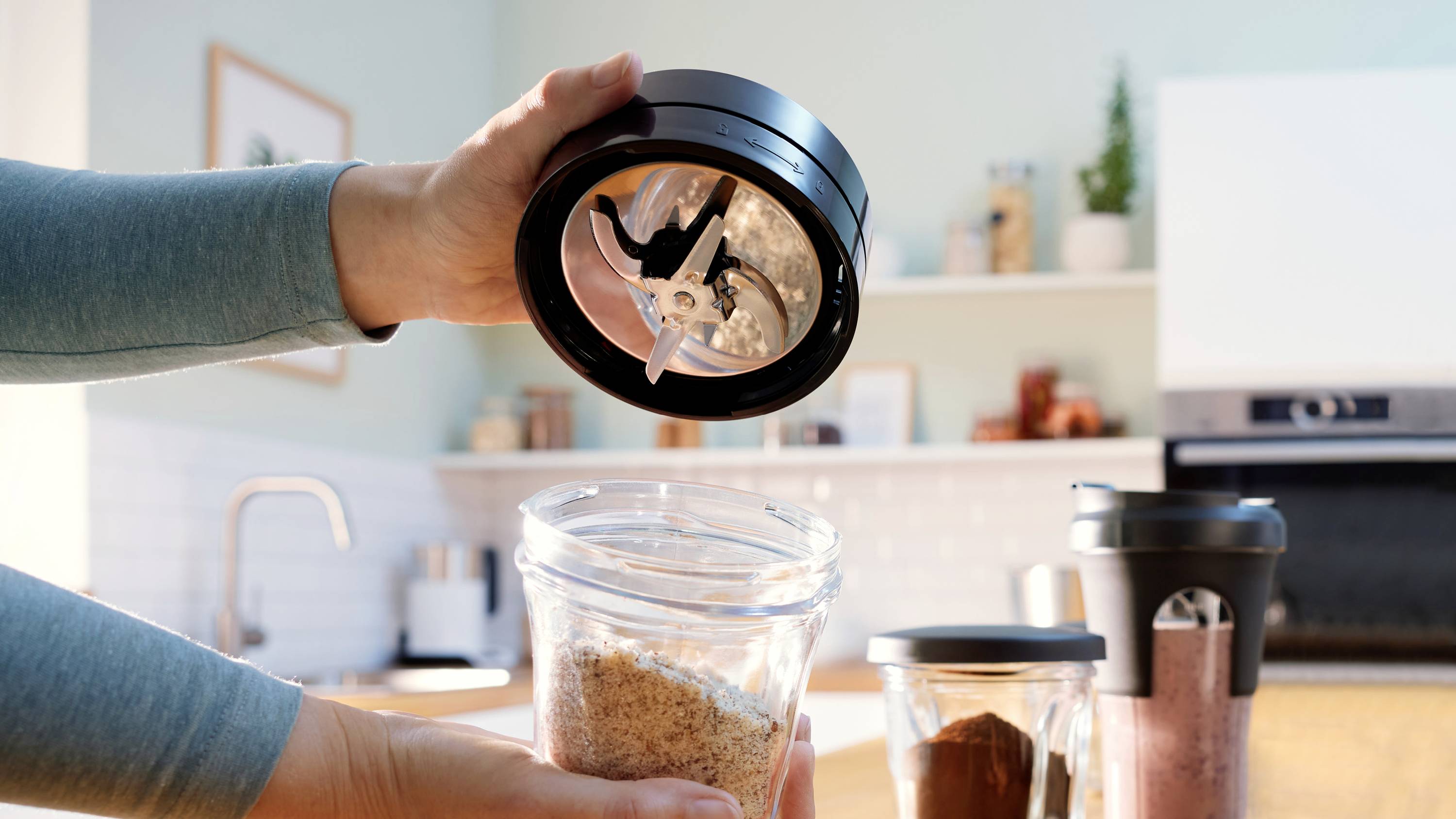 Osoba trzymająca pokrywę blendera z ostrzami nad szklanym pojemnikiem wypełnionym zmielona kawą w kuchni, szykująca się do zmiksowania.