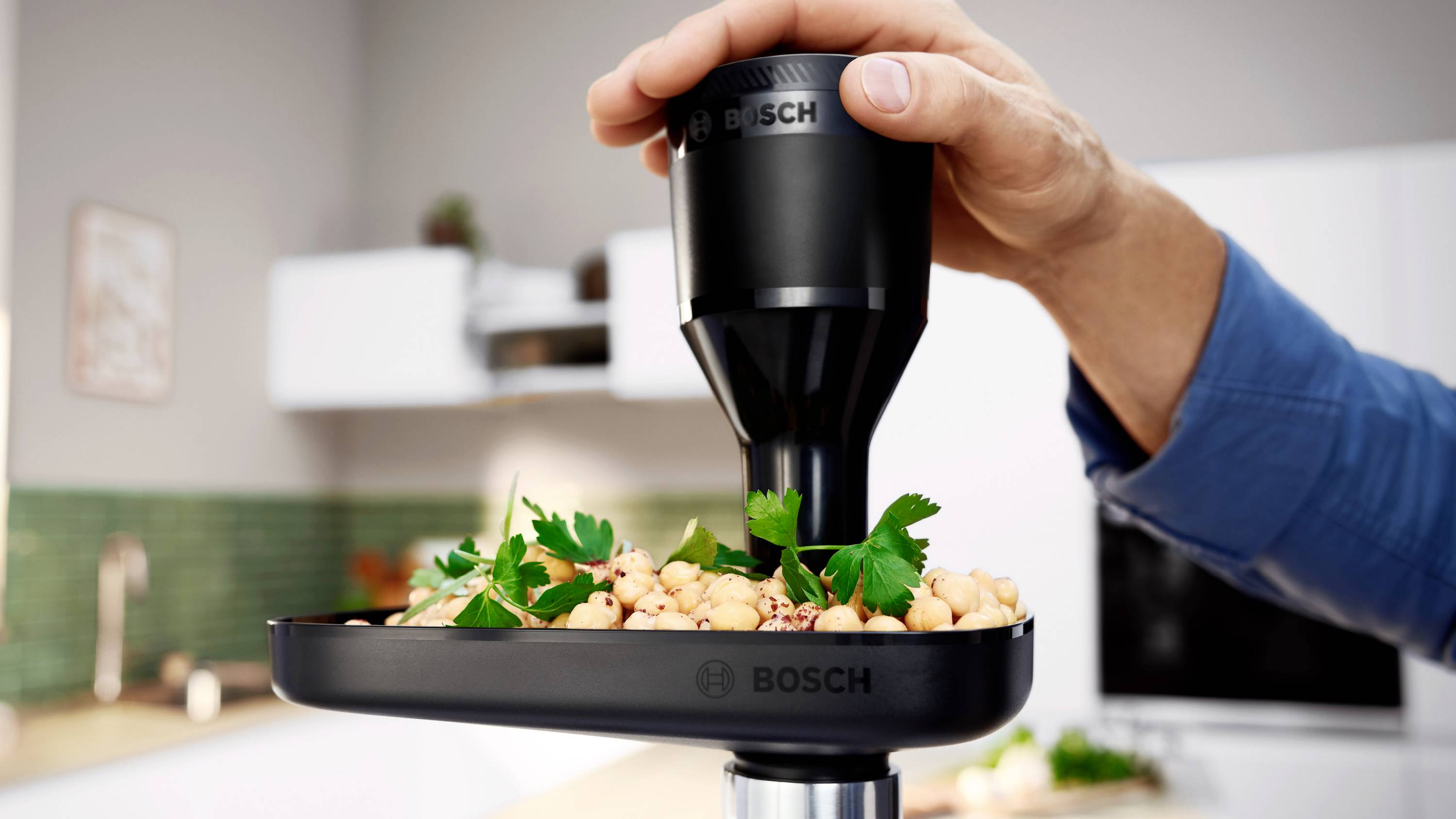 Osoba trzyma ręczny blender Bosch nad miską wypełnioną ciecierzycą i pietruszką, w nowoczesnym wnętrzu kuchennym.