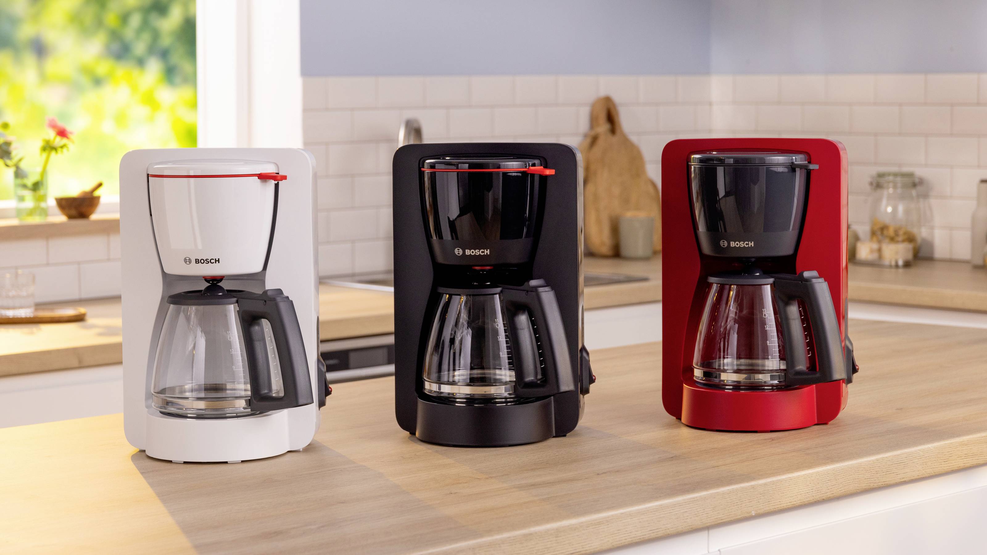 Trzy ekspresy do kawy w kolorach białym, czarnym i czerwonym stoją na blacie kuchennym. Jasna kuchnia z światłem słonecznym w tle.