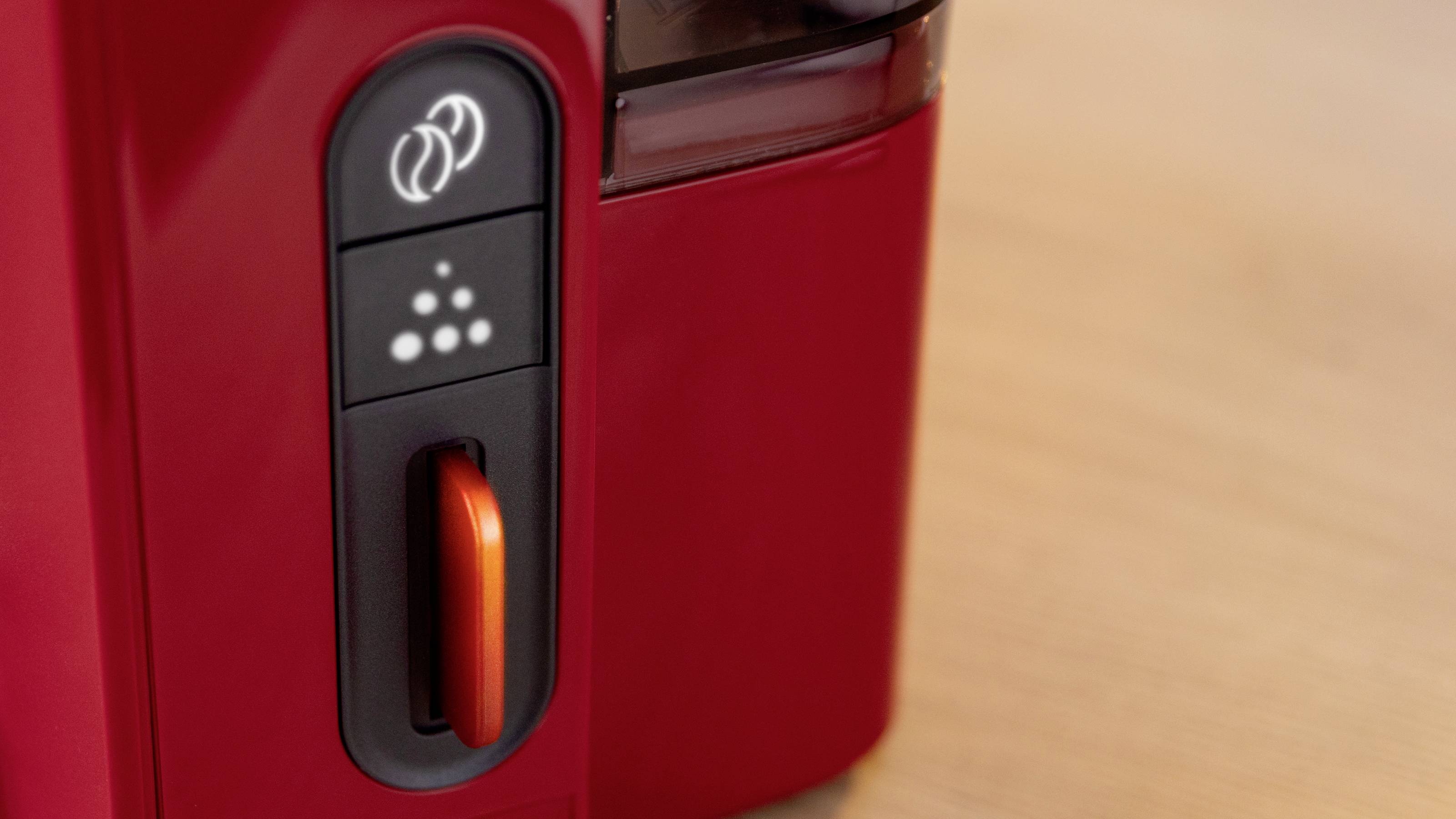 Zbliżenie na czerwony panel sterowania ekspresu do kawy, prezentujący przyciski i pokrętło. Panel zawiera ikony symbolizujące różne opcje parzenia.