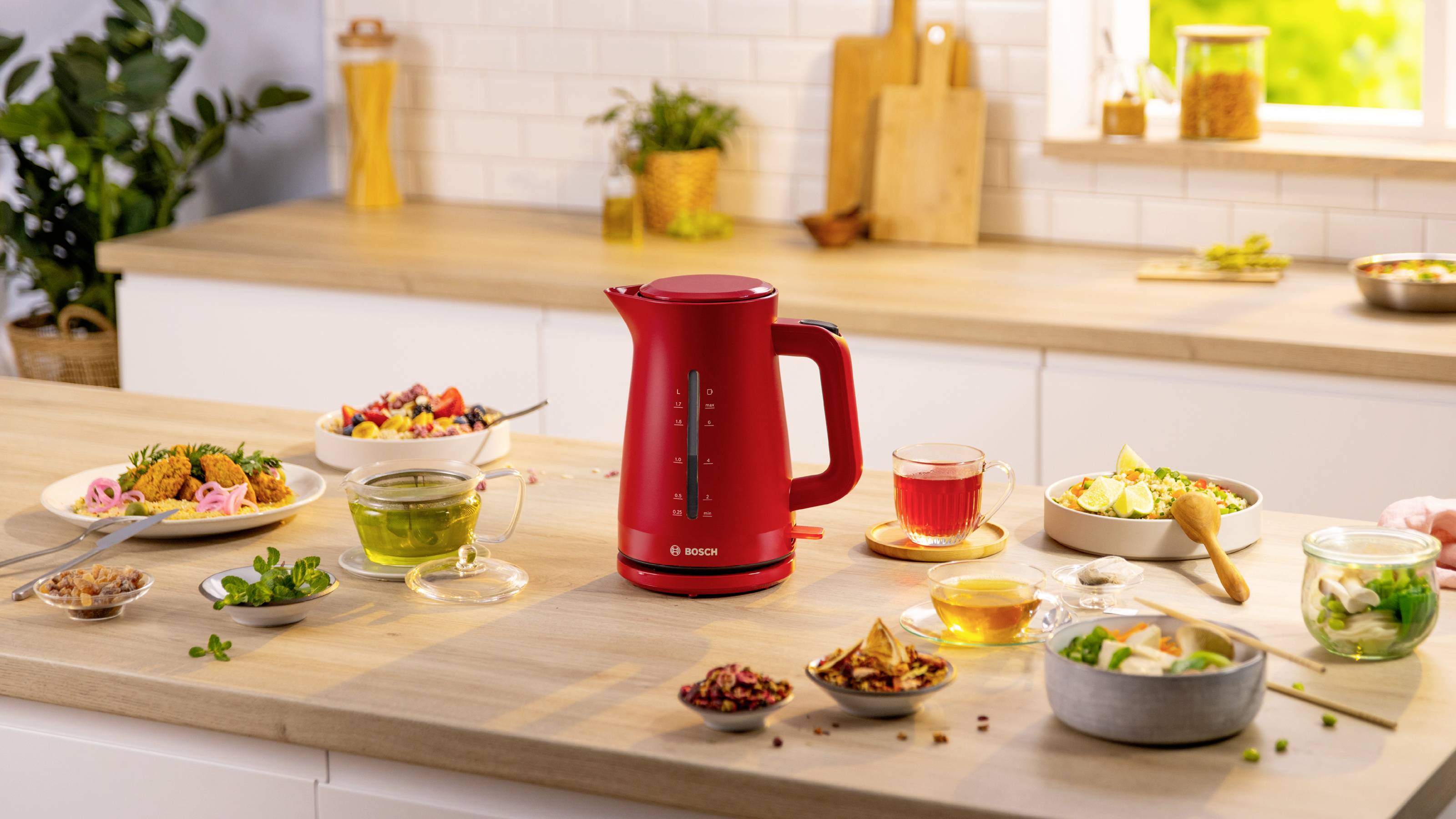 Czerwony elektryczny czajnik stoi na blacie kuchennym, otoczony różnymi naczyniami i napojami, a w tle widać jasne okno i roślinę.