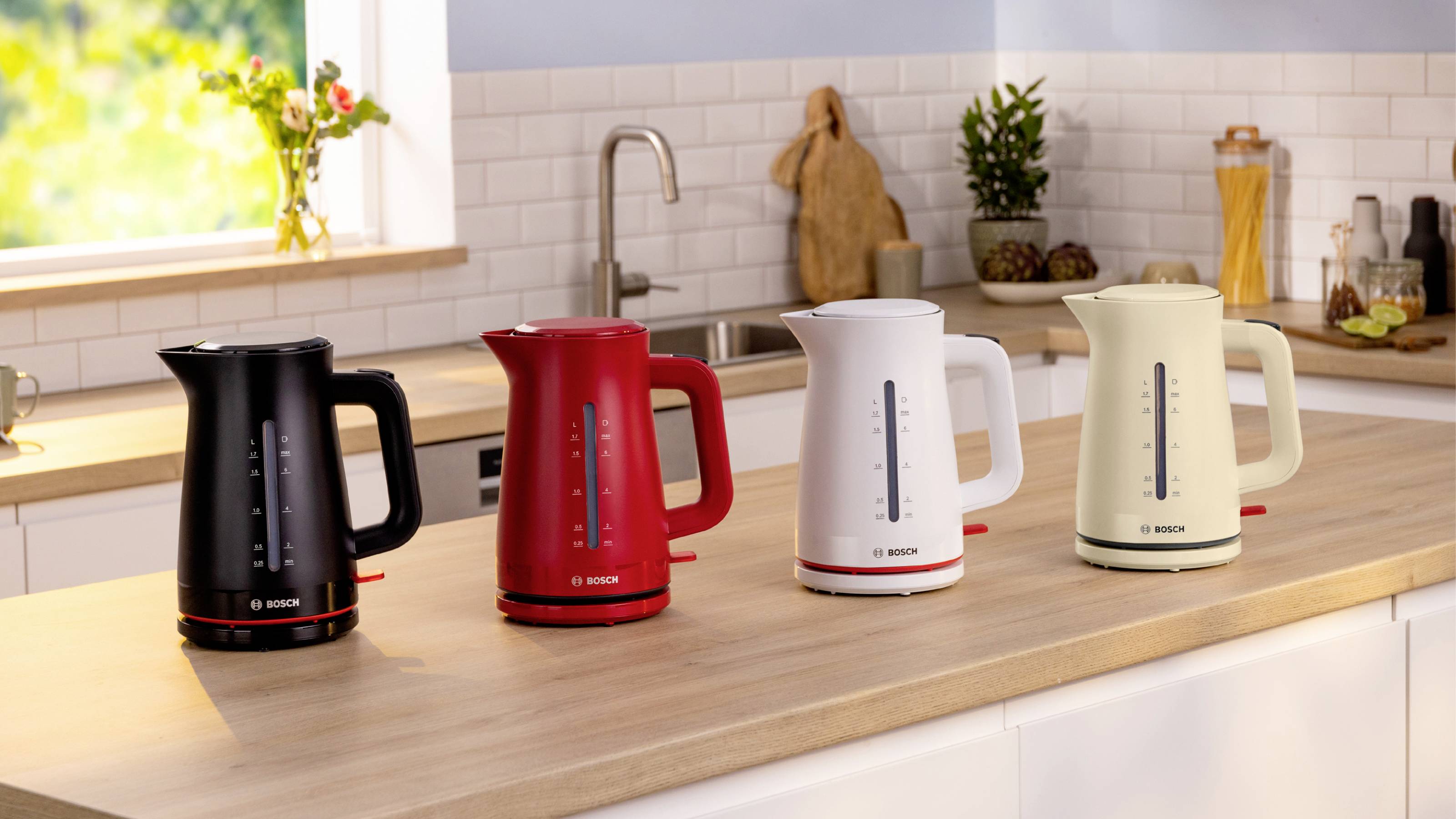 Cztery czajniki elektryczne w kolorach czarnym, czerwonym, białym i kremowym stoją obok siebie na blacie kuchennym w nowoczesnym wnętrzu kuchni.