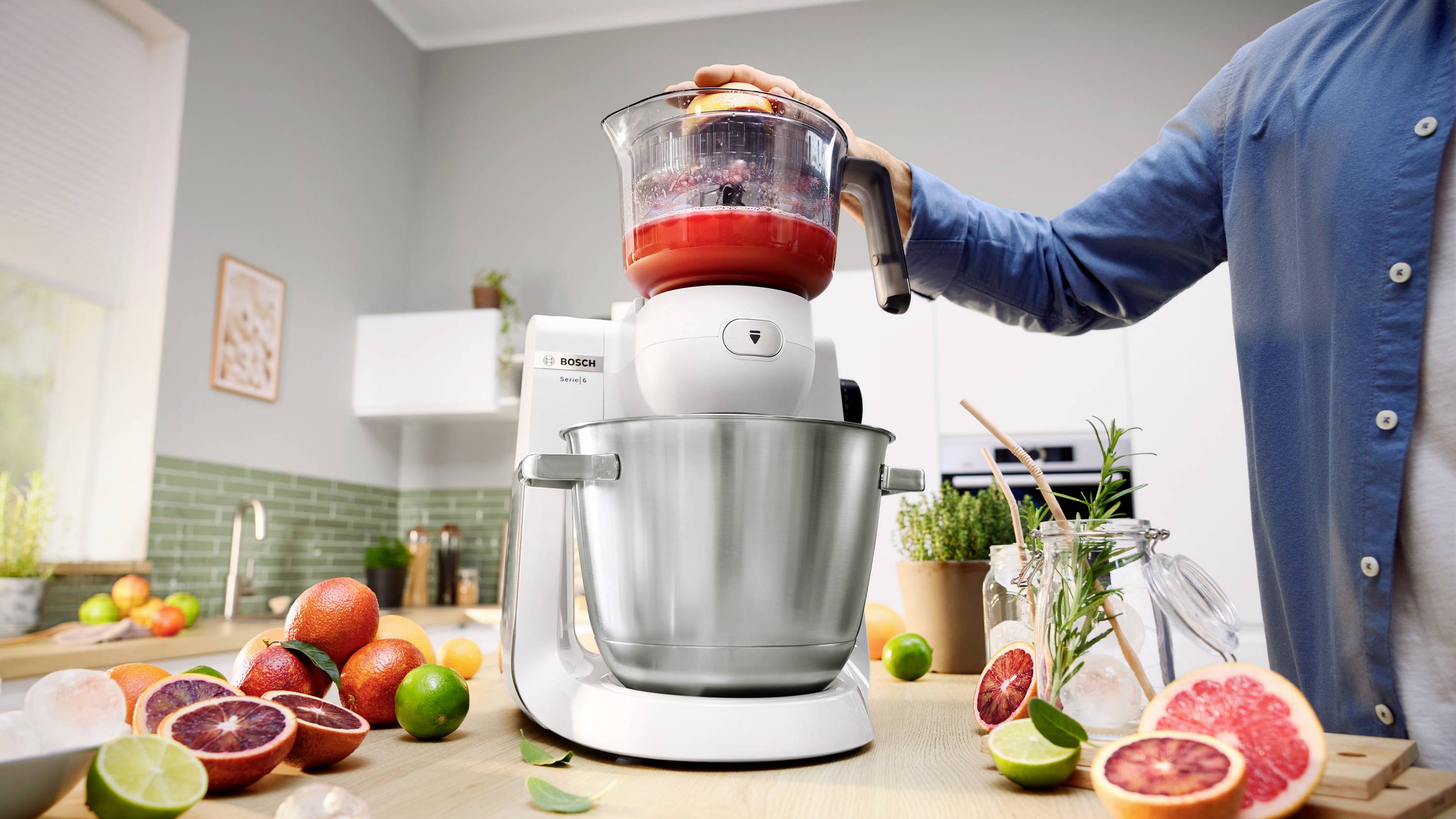 Osoba używa sokowirówki na blacie kuchennym wypełnionym świeżymi owocami cytrusowymi i zielenią, sugerując proces przygotowywania świeżego soku.