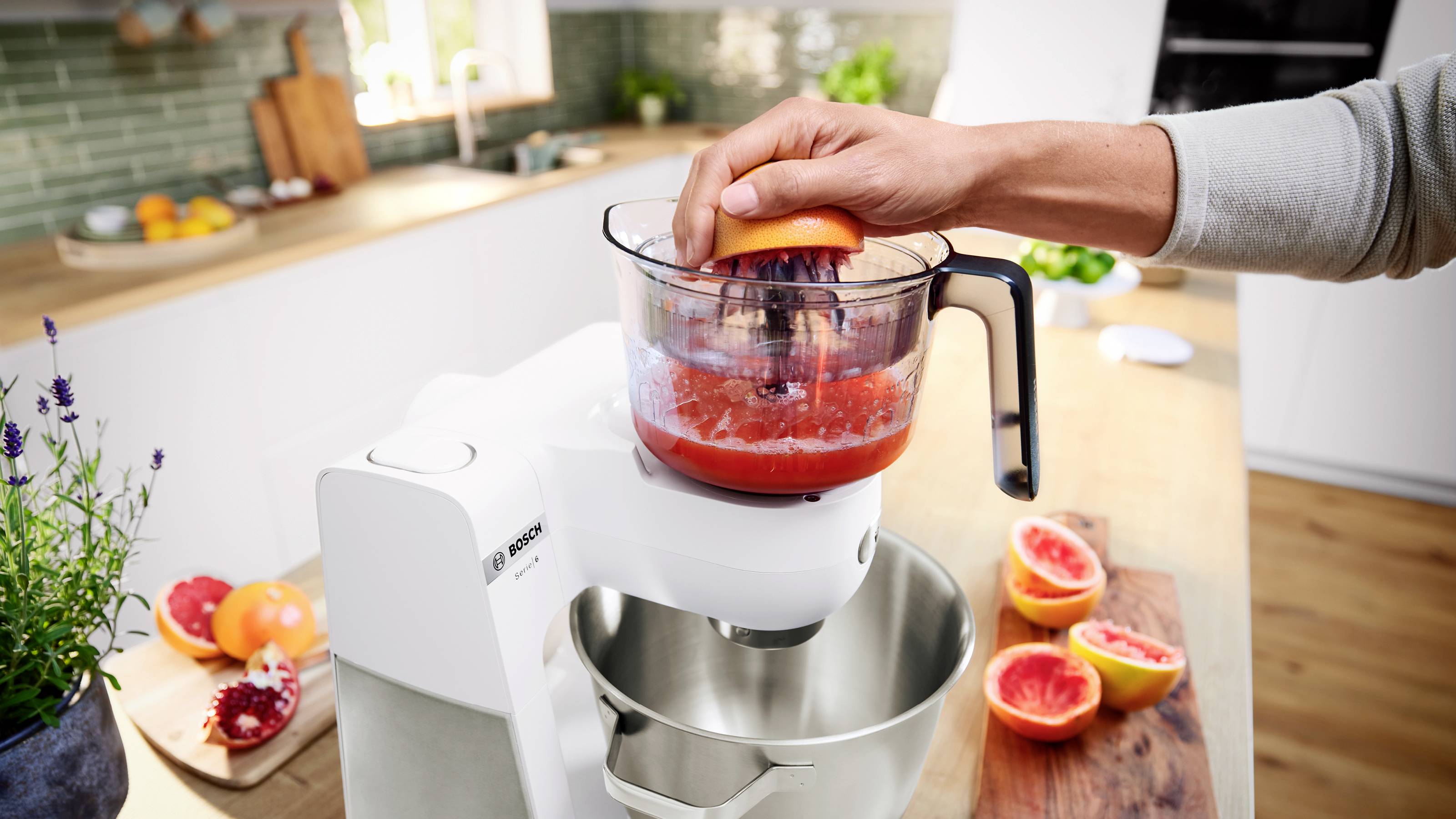 Osoba używa białego miksera elektrycznego do wyciskania grejpfruta, a pocięte owoce widoczne są na drewnianym blacie kuchennym.