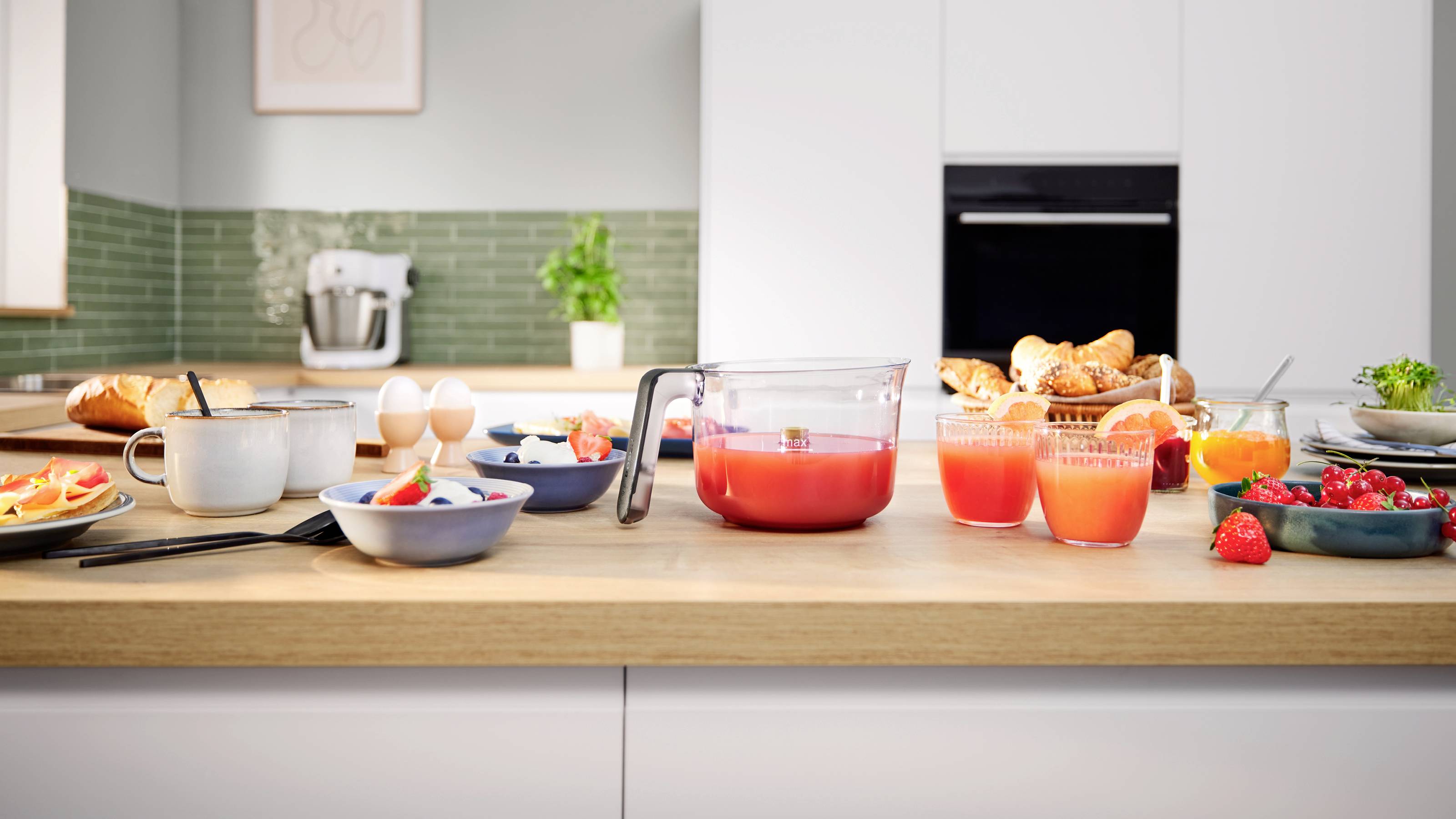 Nowoczesny blat kuchenny z zastawioną do śniadania przestrzenią, na której znajdują się sok, jajka, owoce, chleb i kawa. W tle widoczny jest mikser.