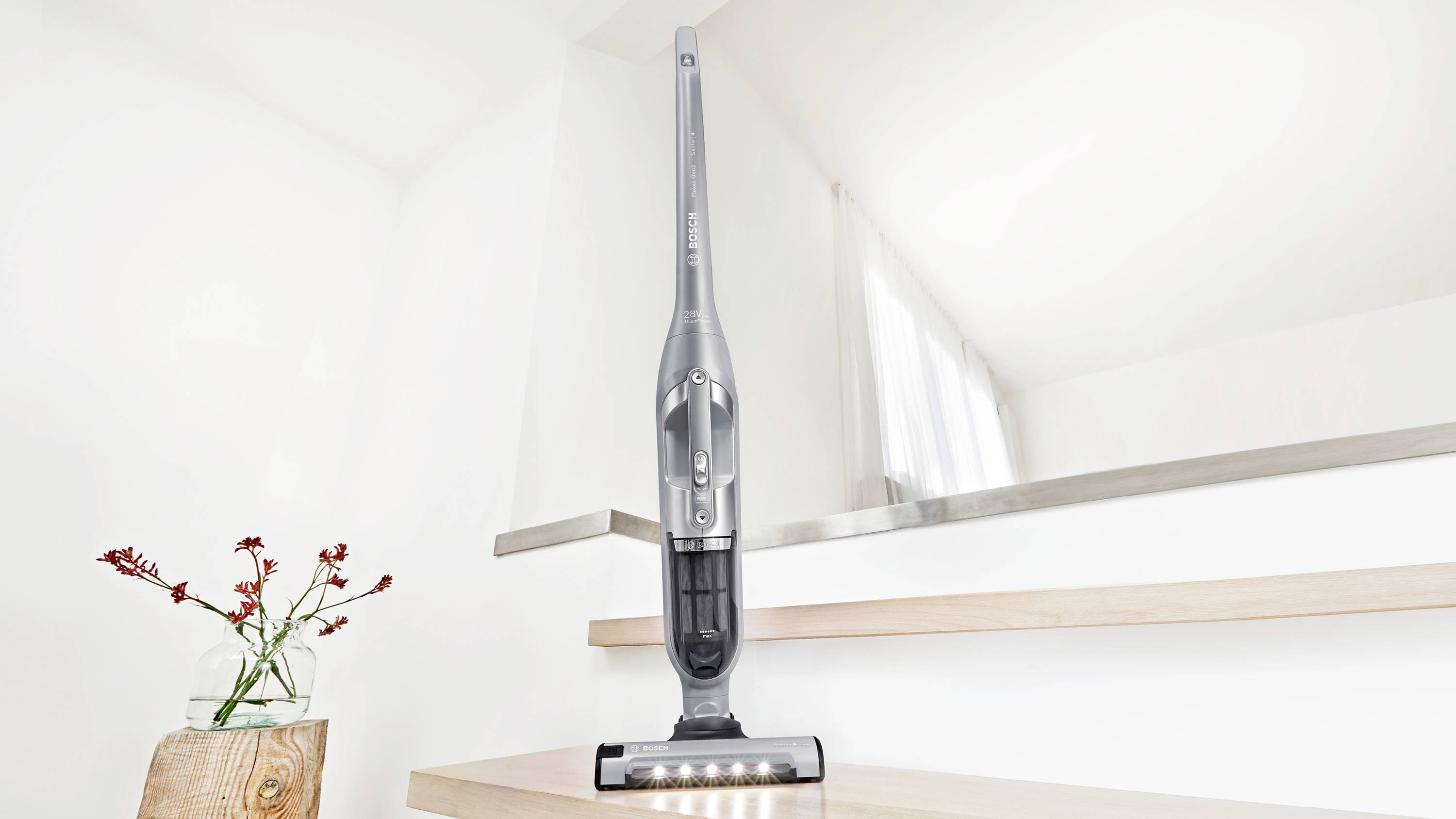 Bezprzewodowy odkurzacz stoi na drewnianych schodach w jasnym pokoju w pobliżu rośliny, podkreślając swój nowoczesny, elegancki wygląd.