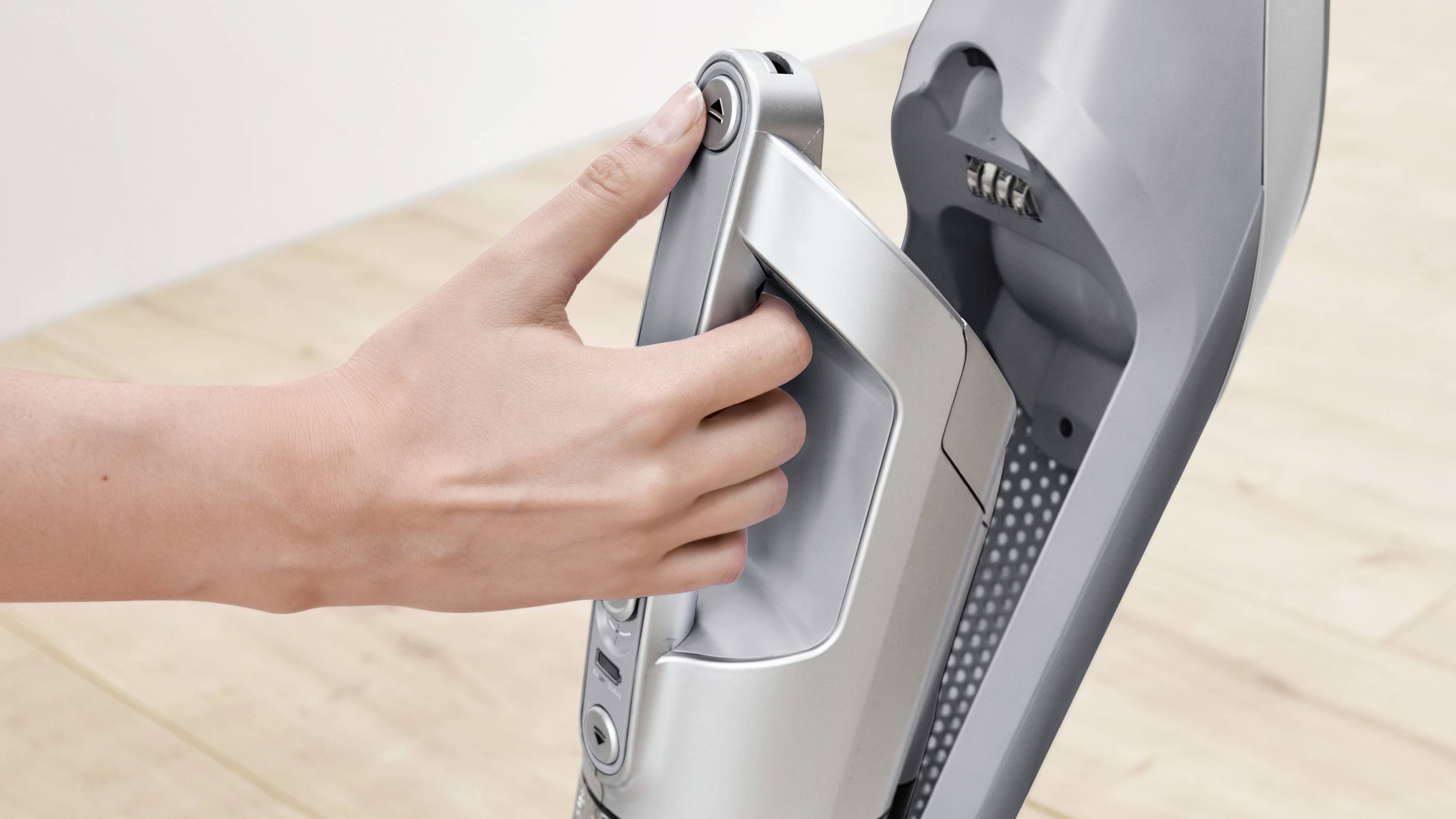 Osoba trzyma uchwyt nowoczesnego, szarego odkurzacza, sugerując łatwe wyjęcie odłączanej części na drewnianej podłodze.