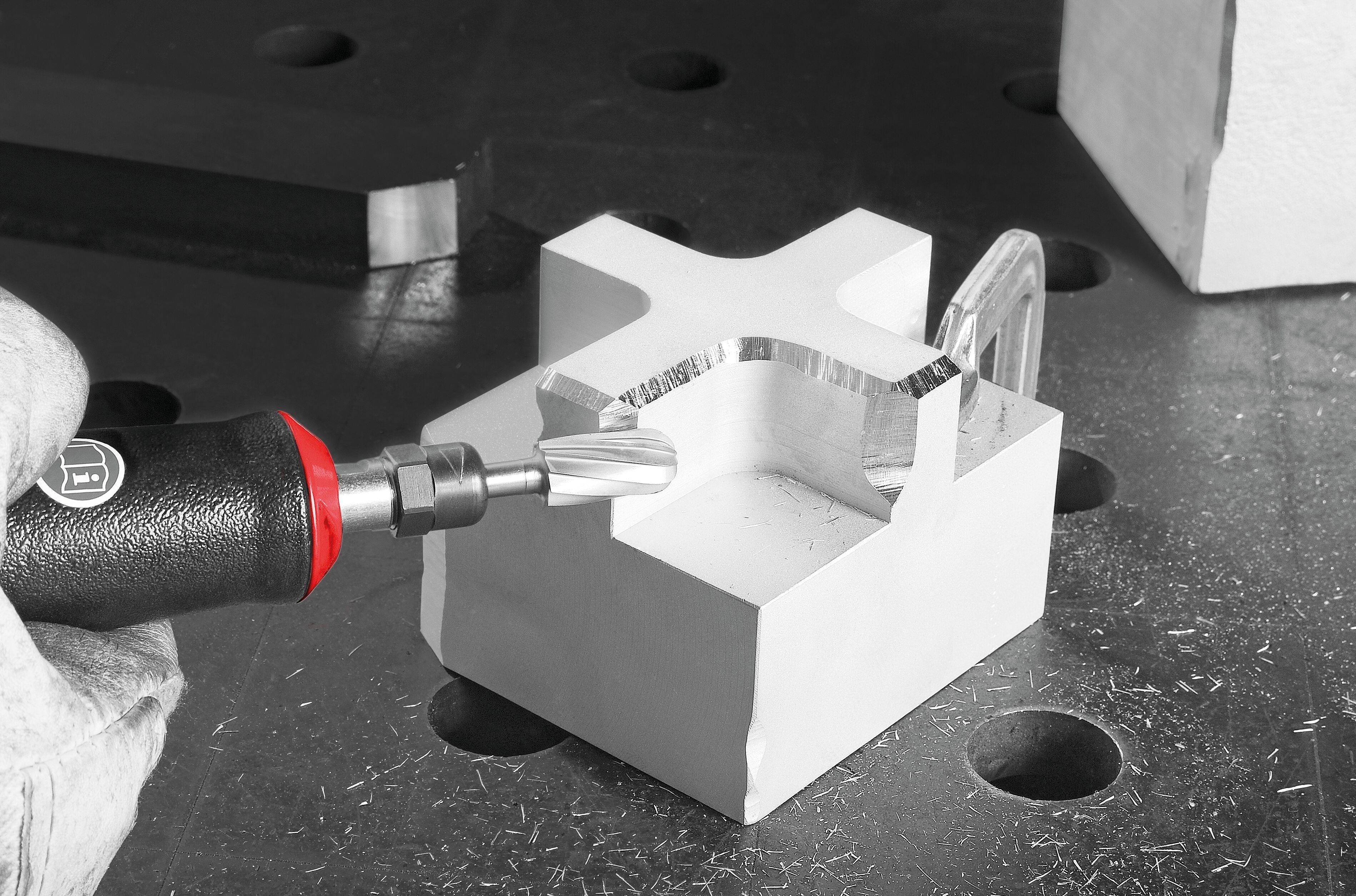 Dłoń w rękawiczce używa czerwono-czarnego narzędzia obrotowego do polerowania krawędzi metalowego, kształtem przypominającego literę X przedmiotu na ciemnej, dziurkowanej powierzchni.