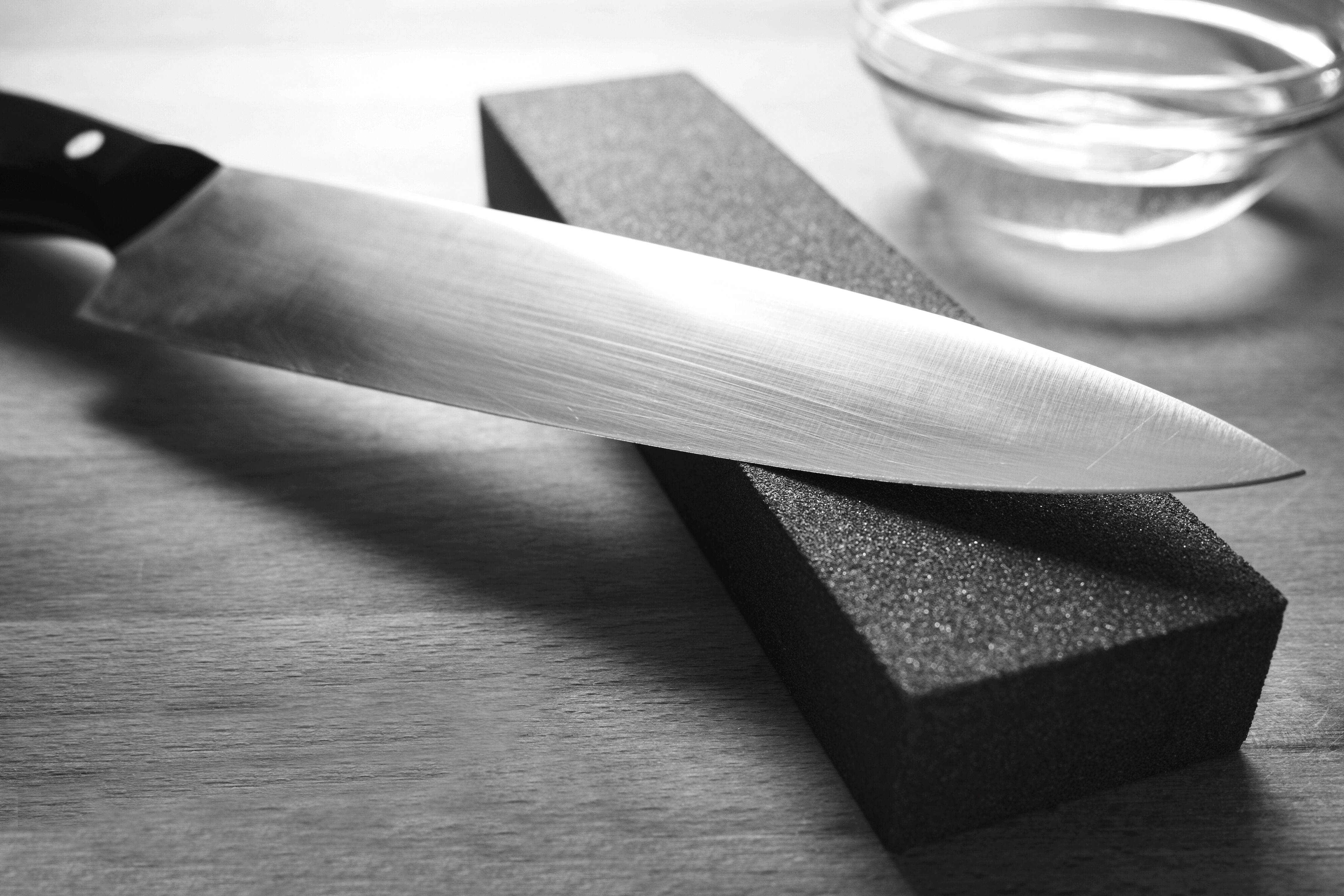 Ostry nóż kuchenny spoczywa na prostokątnym kamieniu do ostrzenia, obok znajduje się mała szklana miska na drewnianej powierzchni, w czerni i bieli.