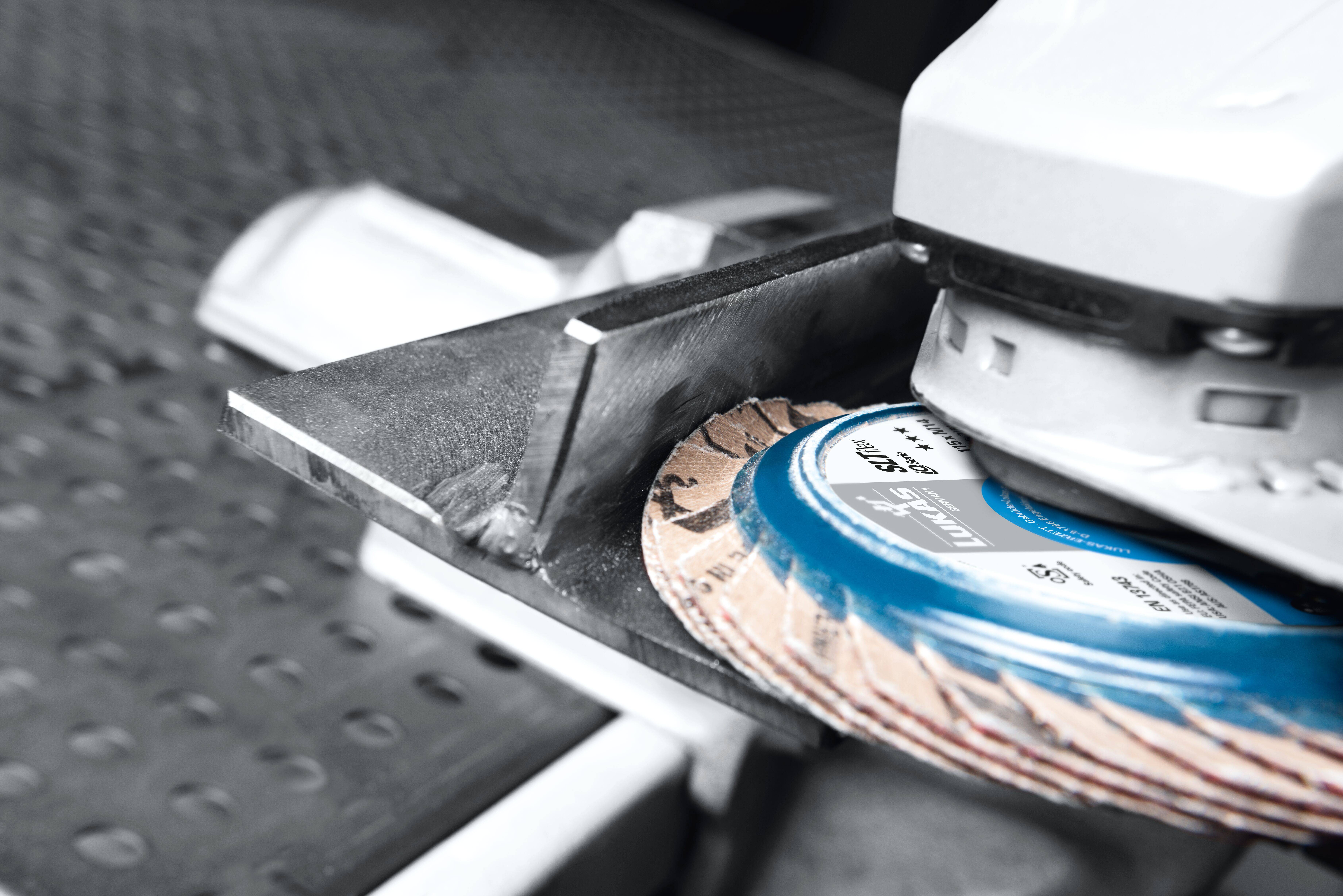 Zbliżenie narzędzia szlifierskiego z niebiesko-brązową tarczą, wygładzającą krawędź arkusza metalu na chropowatej powierzchni stołu warsztatowego.