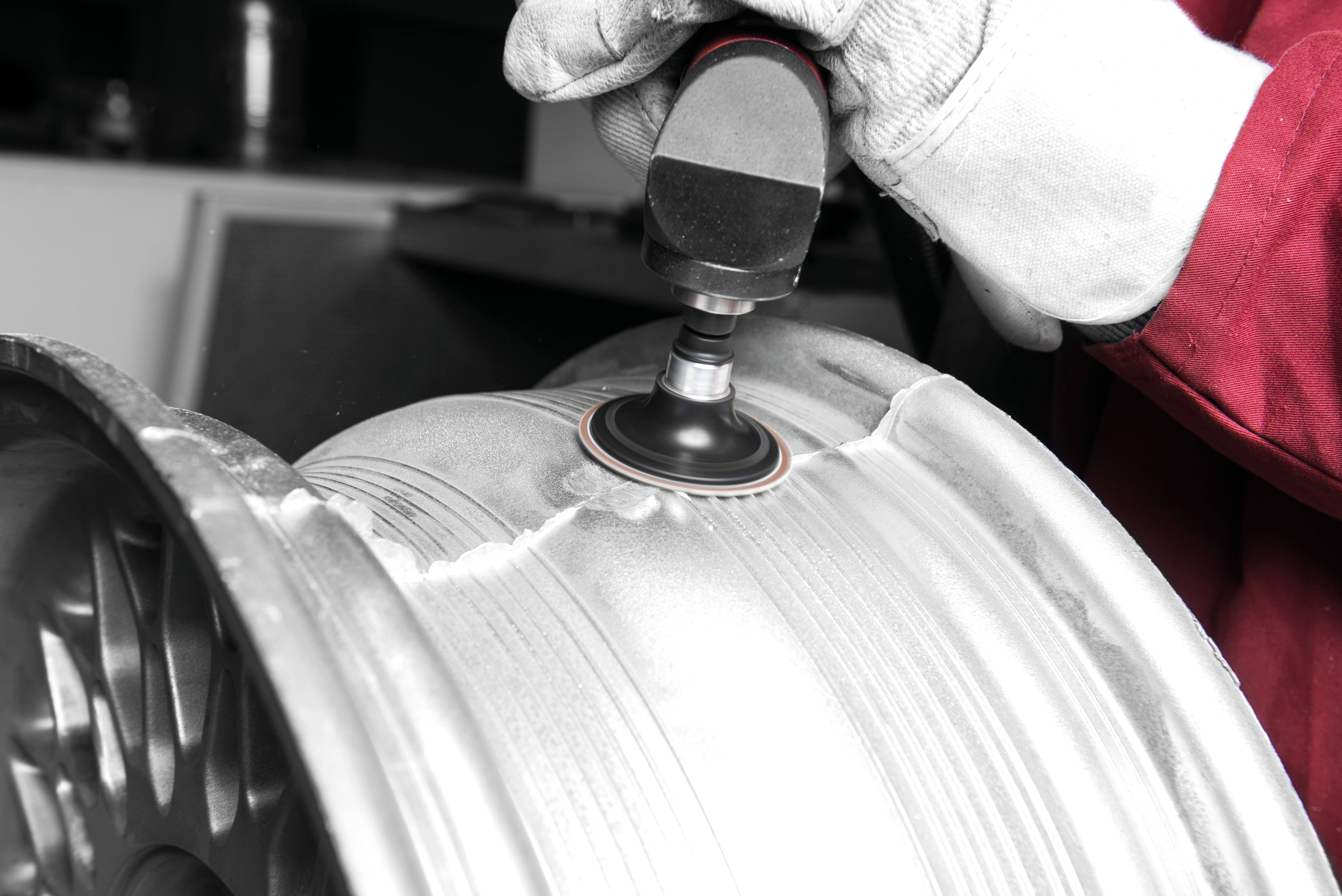 Osoba w rękawicach używa elektronarzędzia do polerowania metalowego felgi samochodowej, koncentrując się na wygładzaniu zadrapań na jej powierzchni.