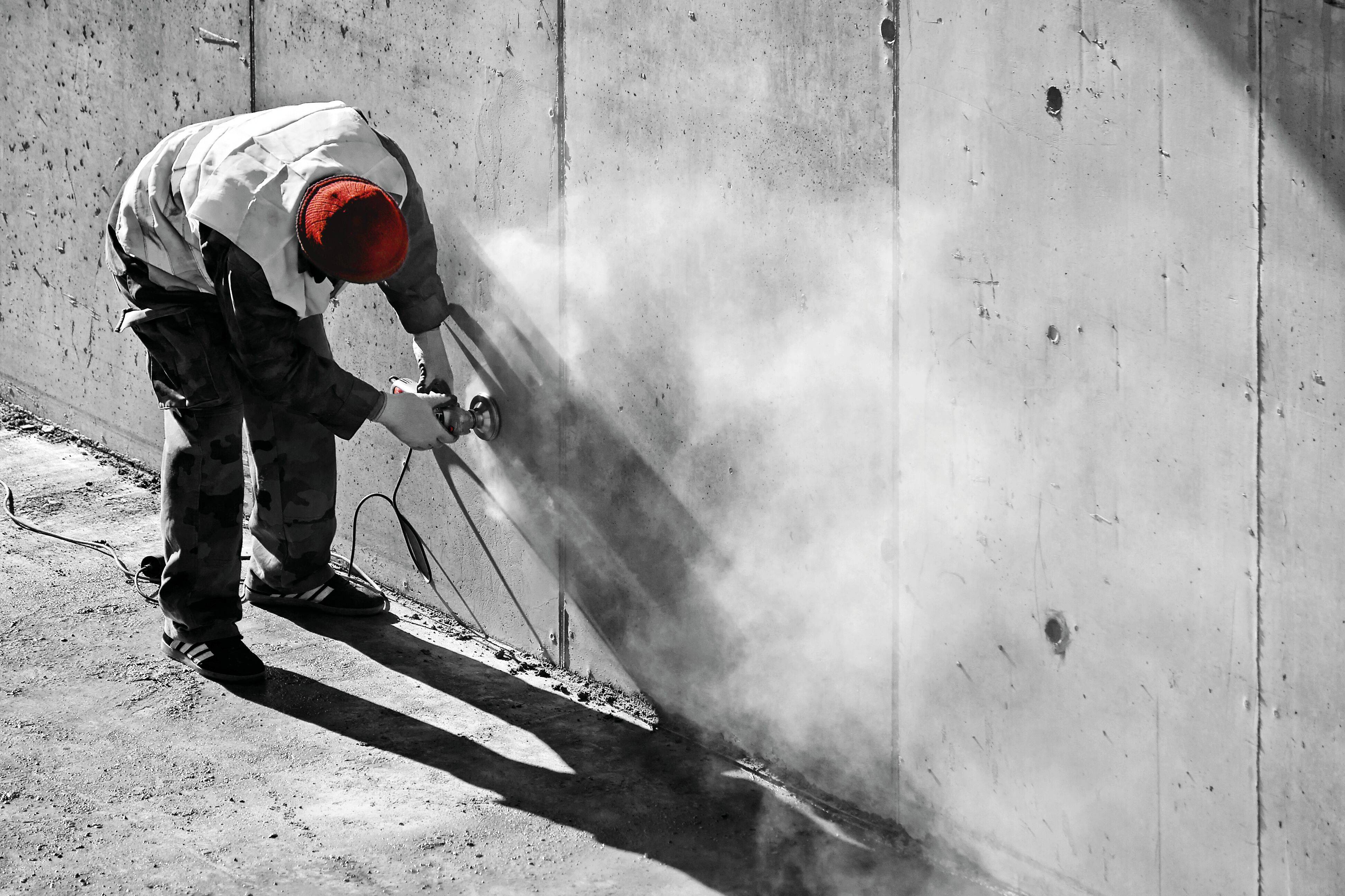 Robotnik używa elektronarzędzia do cięcia betonowej ściany, powodując kłęby pyłu. Ma na głowie czerwoną czapkę i jest ubrany w odzież ochronną.