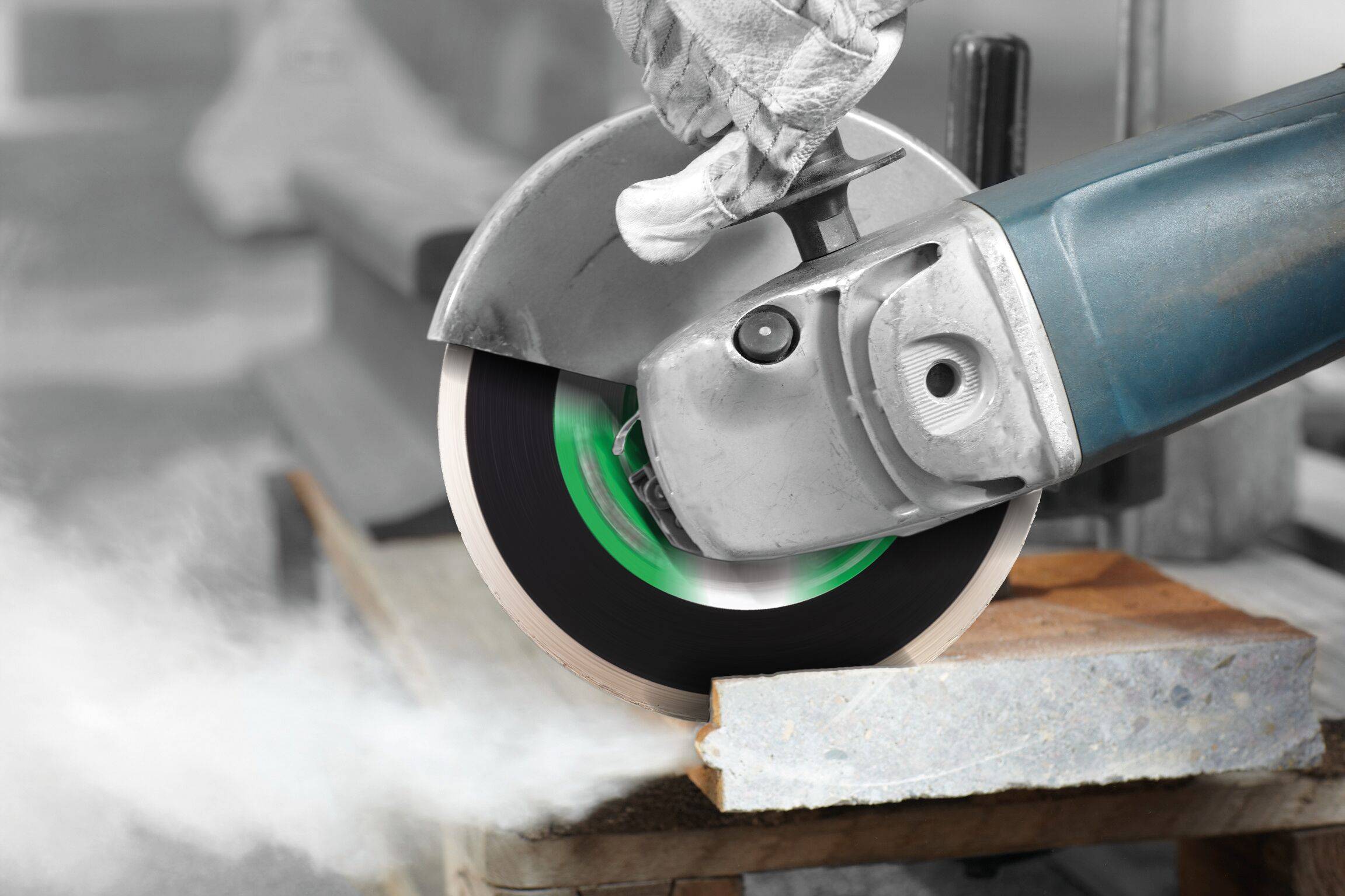 Pracownik używający szlifierki kątowej z zieloną tarczą tnie płytę kamienną, wytwarzając widoczny pył. Pracownik nosi rękawice ochronne.