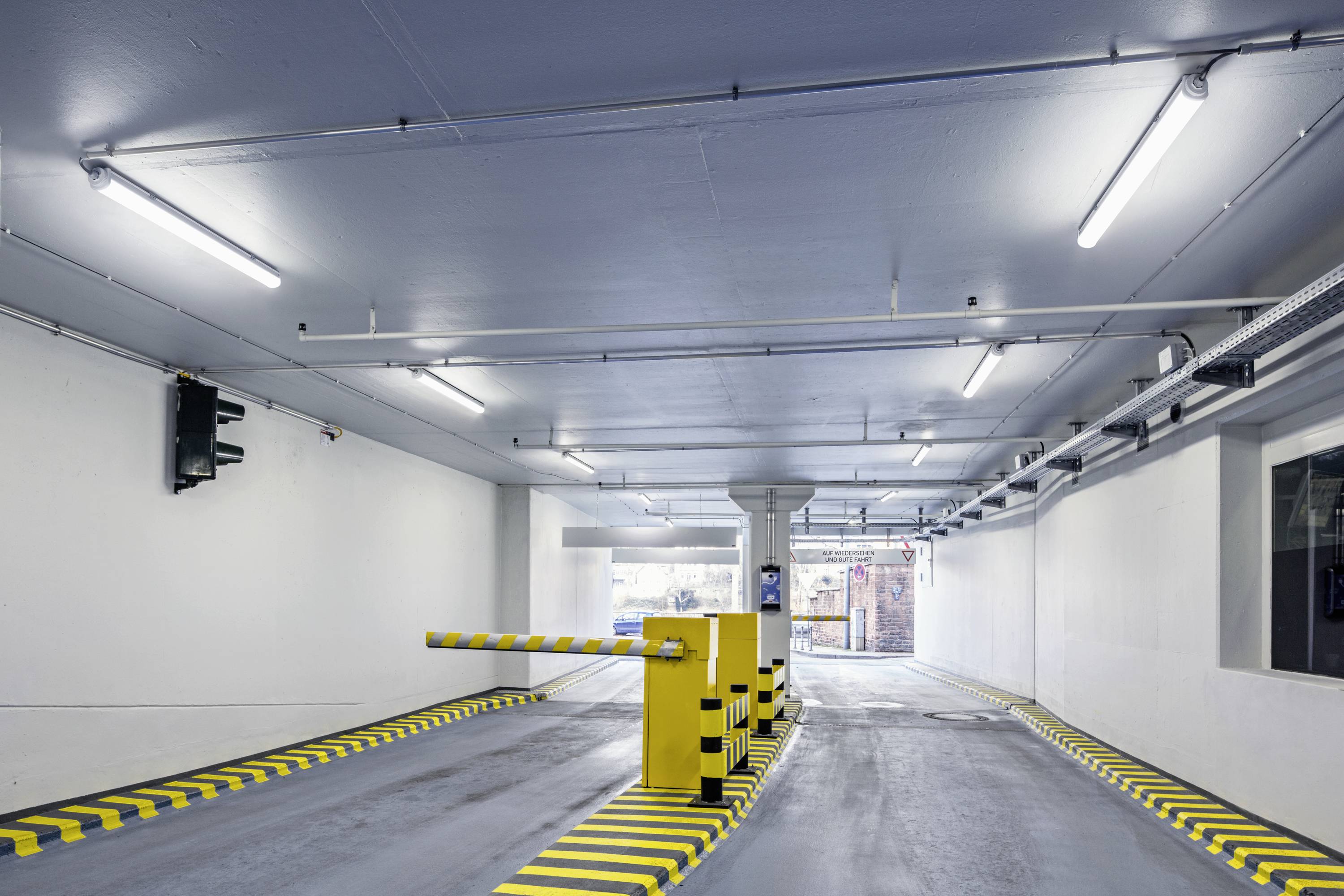 Dobrze oświetlone wejście do parkingu wielopoziomowego z żółto-czarną zaporą blokującą przejazd oraz kamerami bezpieczeństwa zamontowanymi na ścianach.