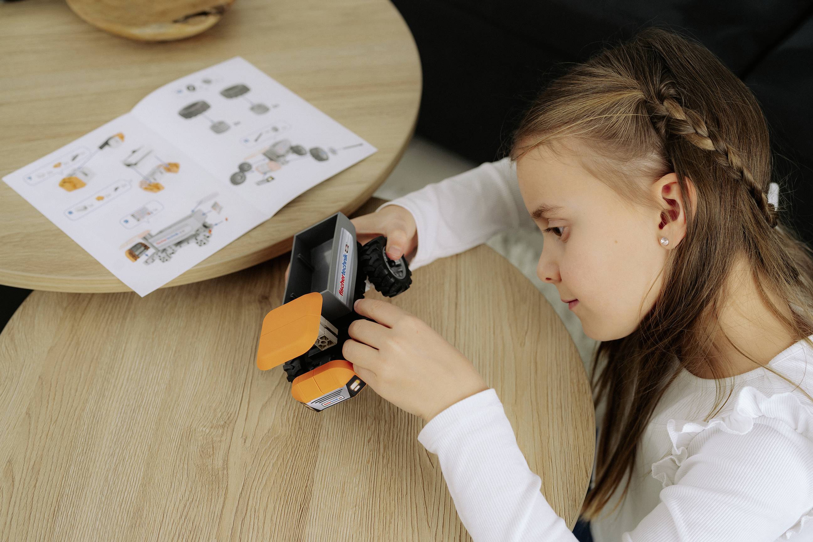 Młoda dziewczynka montuje mały pojazd robotyczny, korzystając z instrukcji leżących na stole, jest całkowicie skupiona i zaangażowana, co podkreśla naukę w dziedzinie STEM.