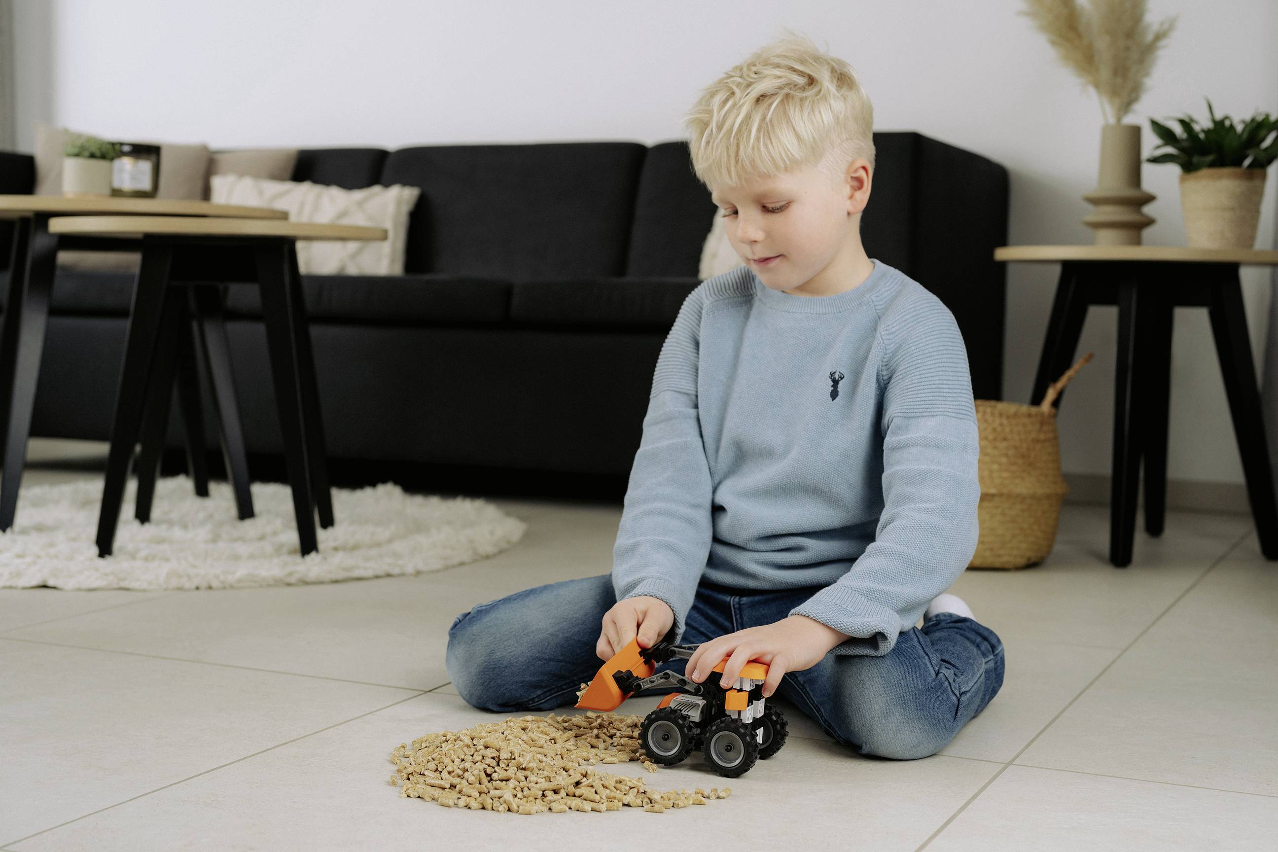 Dziecko o włosach blond siedzące na podłodze w salonie, bawiące się zabawkową koparką i drewnianymi klockami.