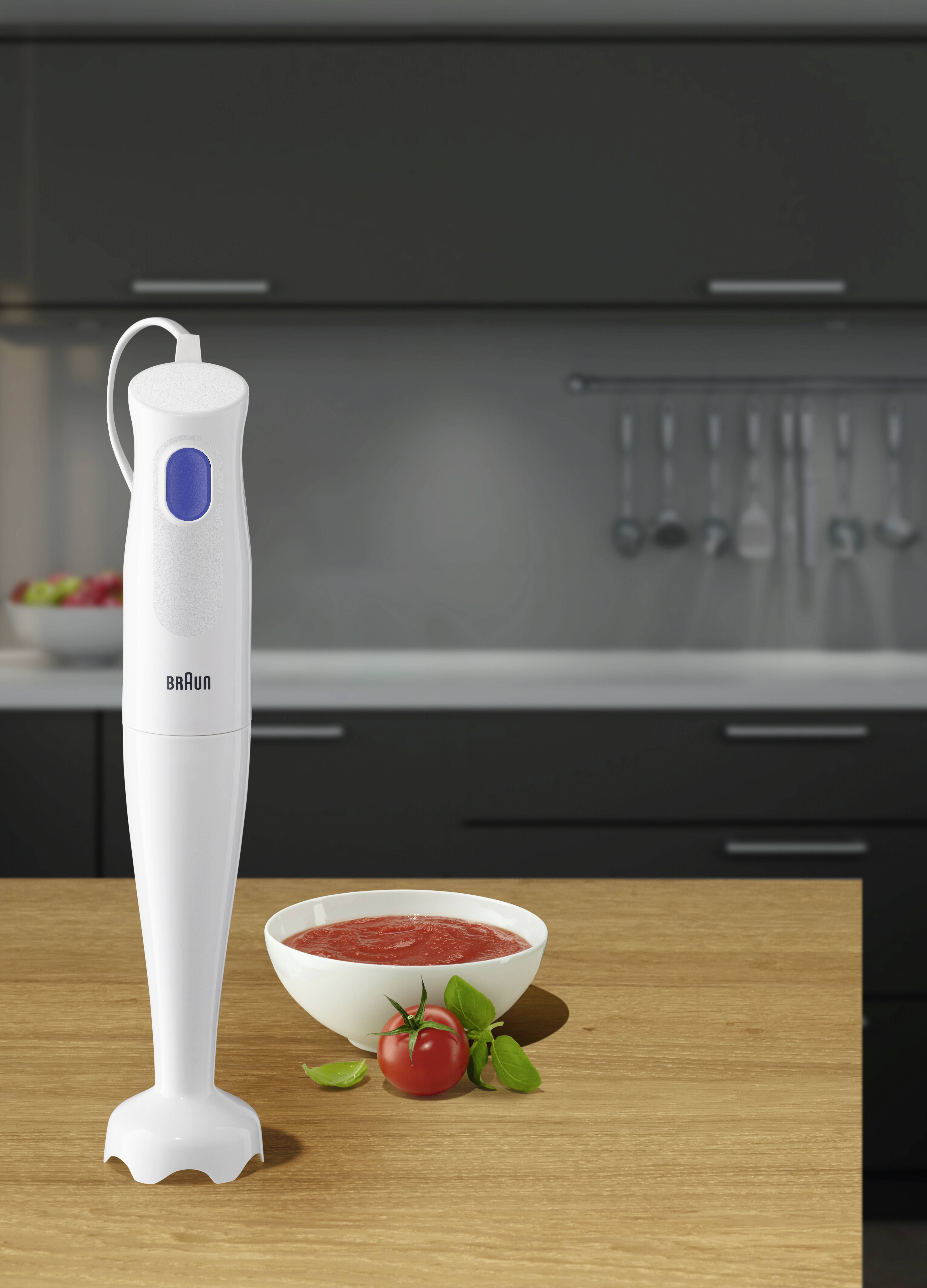 Biały blender ręczny na blacie kuchennym obok miski zupy pomidorowej i świeżego pomidora, na tle nowoczesnej kuchni.