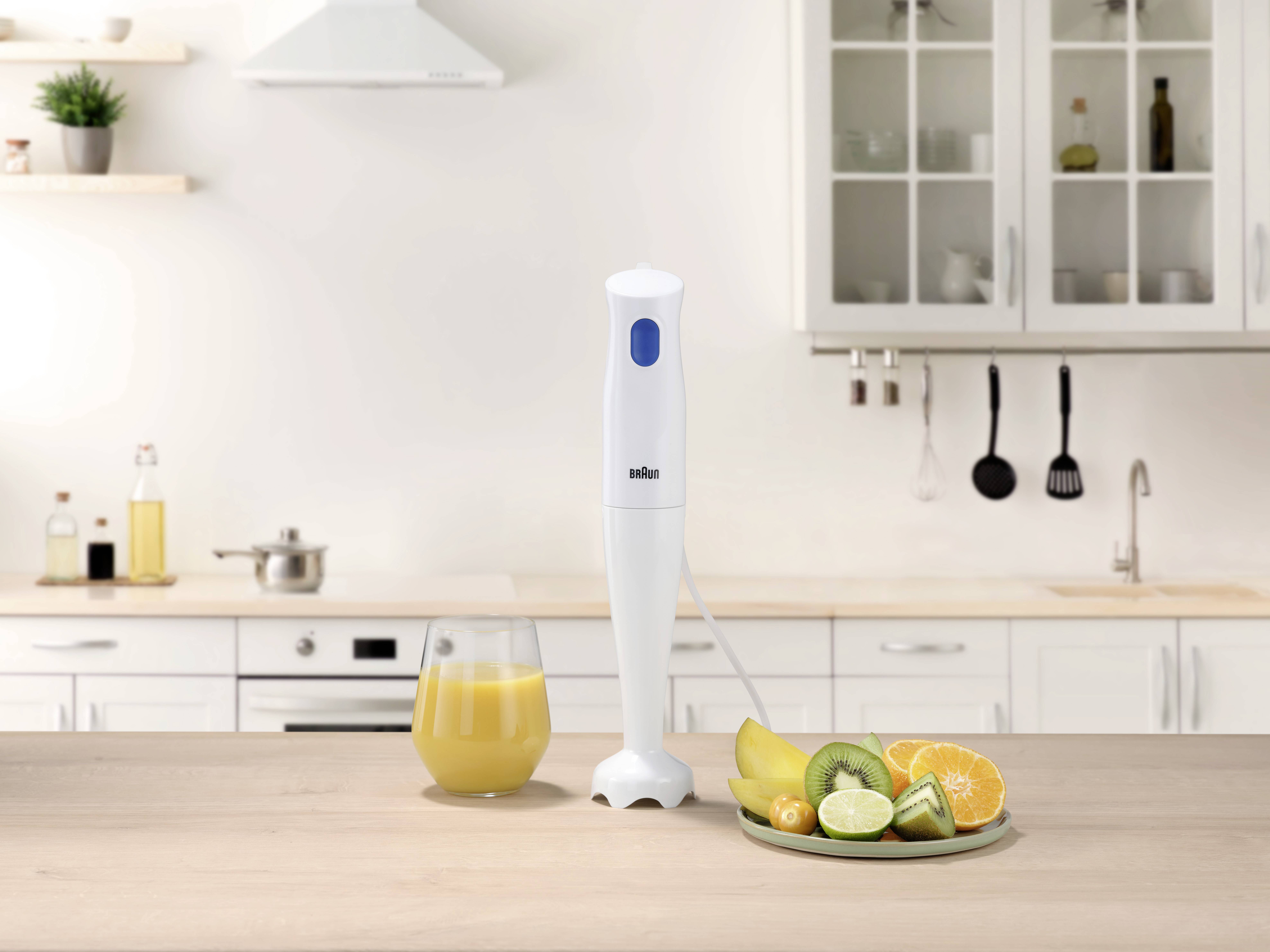 Biały blender ręczny stoi na blacie kuchennym obok szklanki żółtego soku oraz talerza z pokrojonymi owocami, w tym pomarańczami i kiwi.