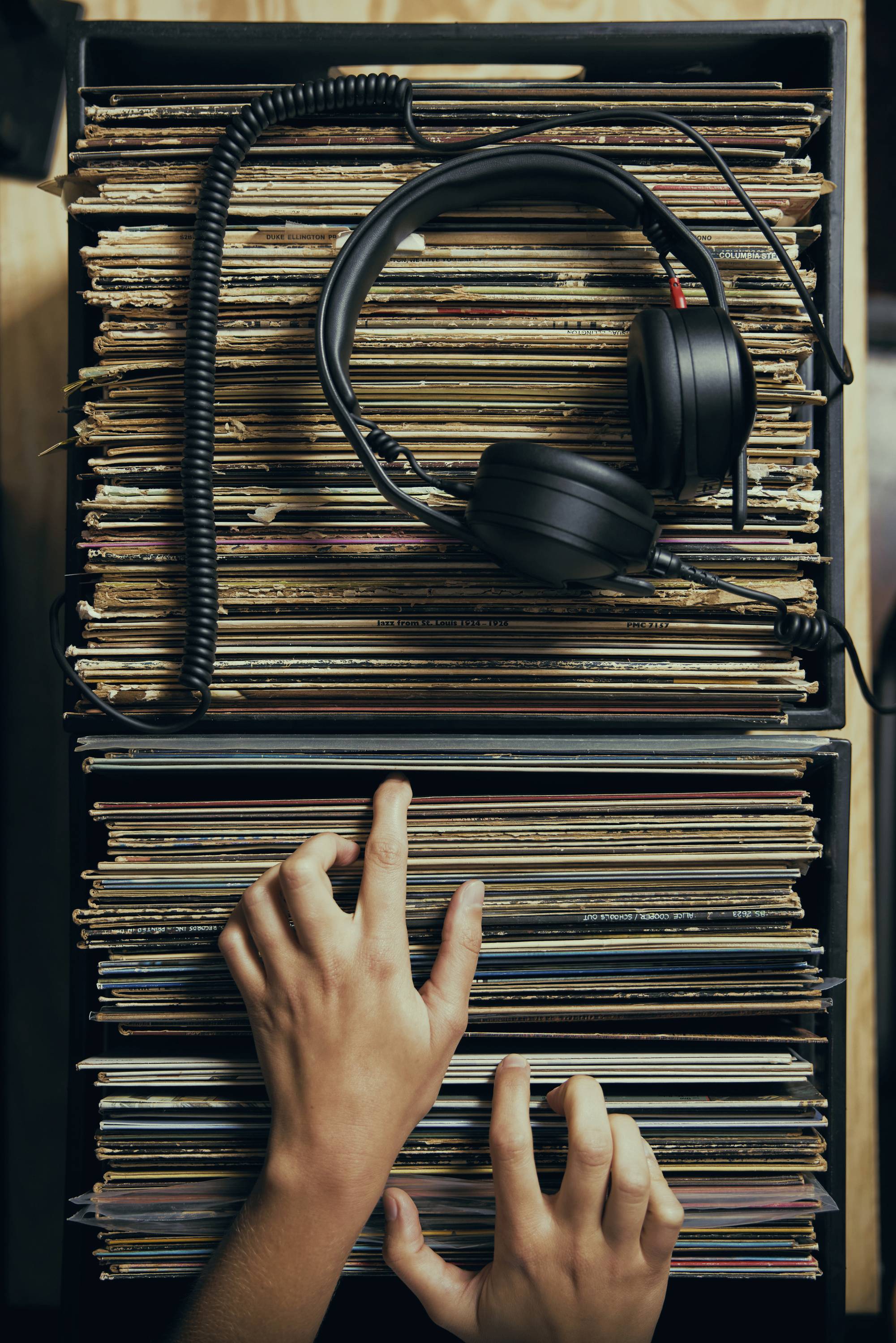 Para rąk przebierających przez kolekcję płyt winylowych w pudełku, z parą słuchawek spoczywających na wierzchu.