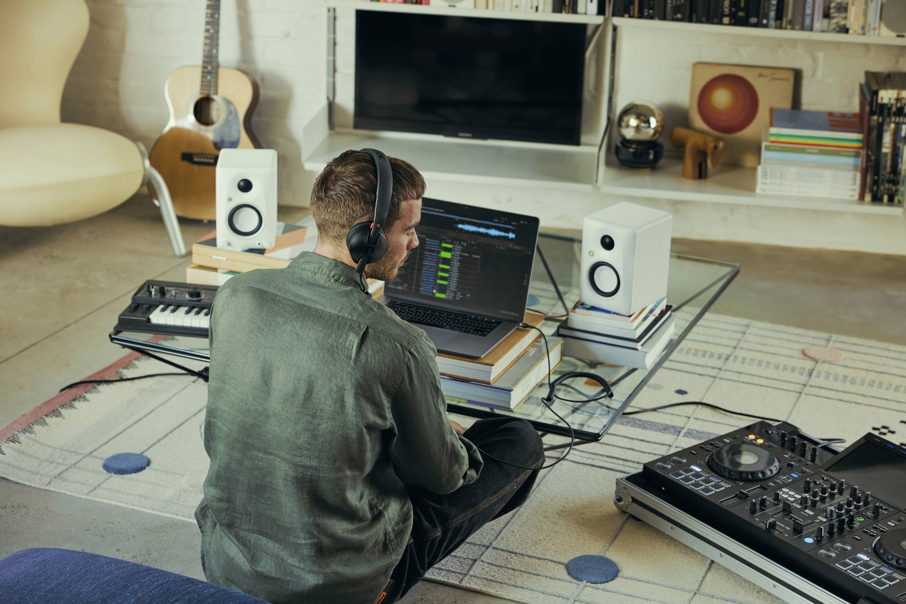Osoba w słuchawkach siedzi na podłodze, pracując na laptopie z oprogramowaniem do produkcji muzycznej. Wokół stanowiska znajduje się sprzęt muzyczny.