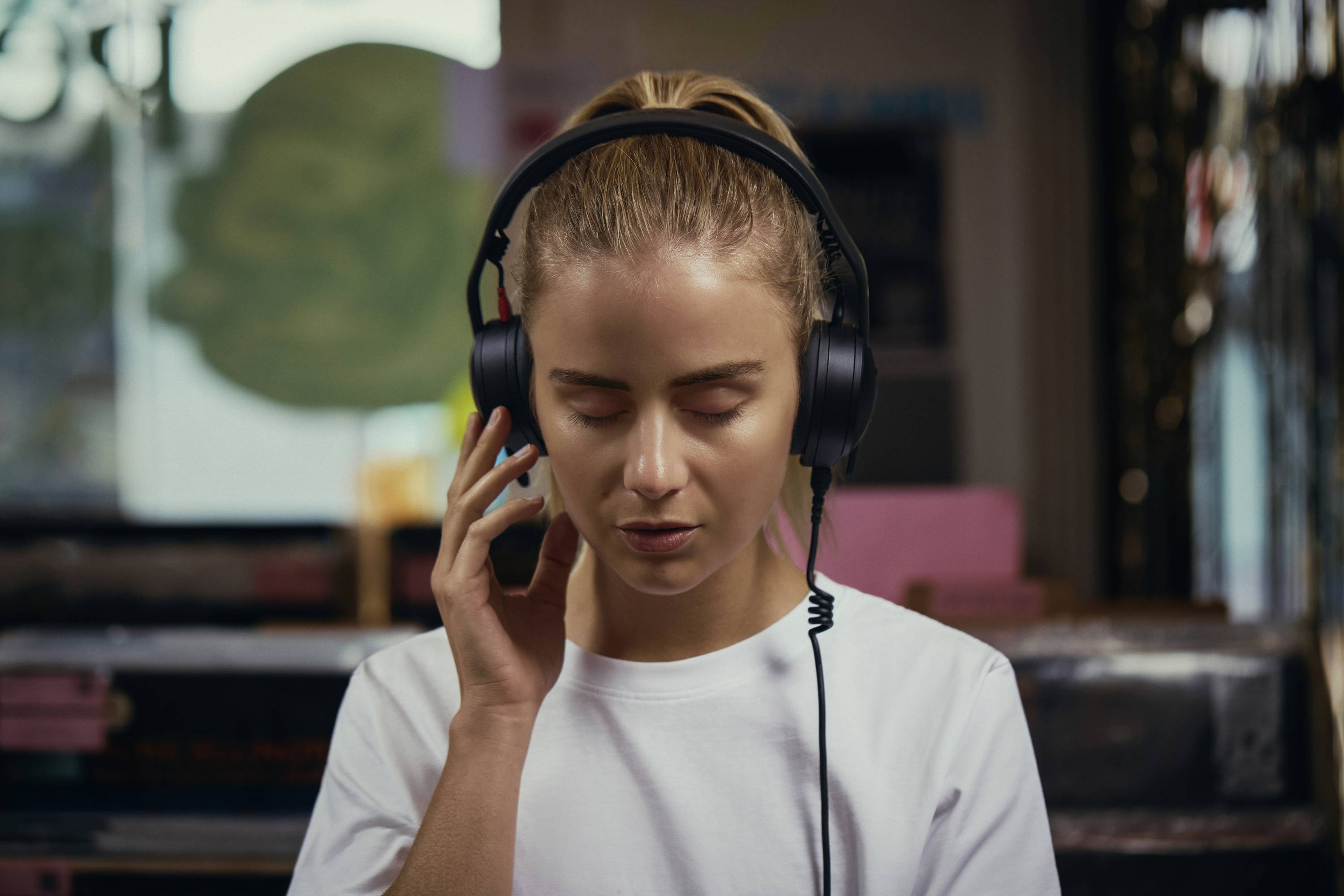 Osoba z zamkniętymi oczami słucha muzyki na słuchawkach, stojąc w pomieszczeniu z rozmytym tłem, wyrażająca stan relaksu.