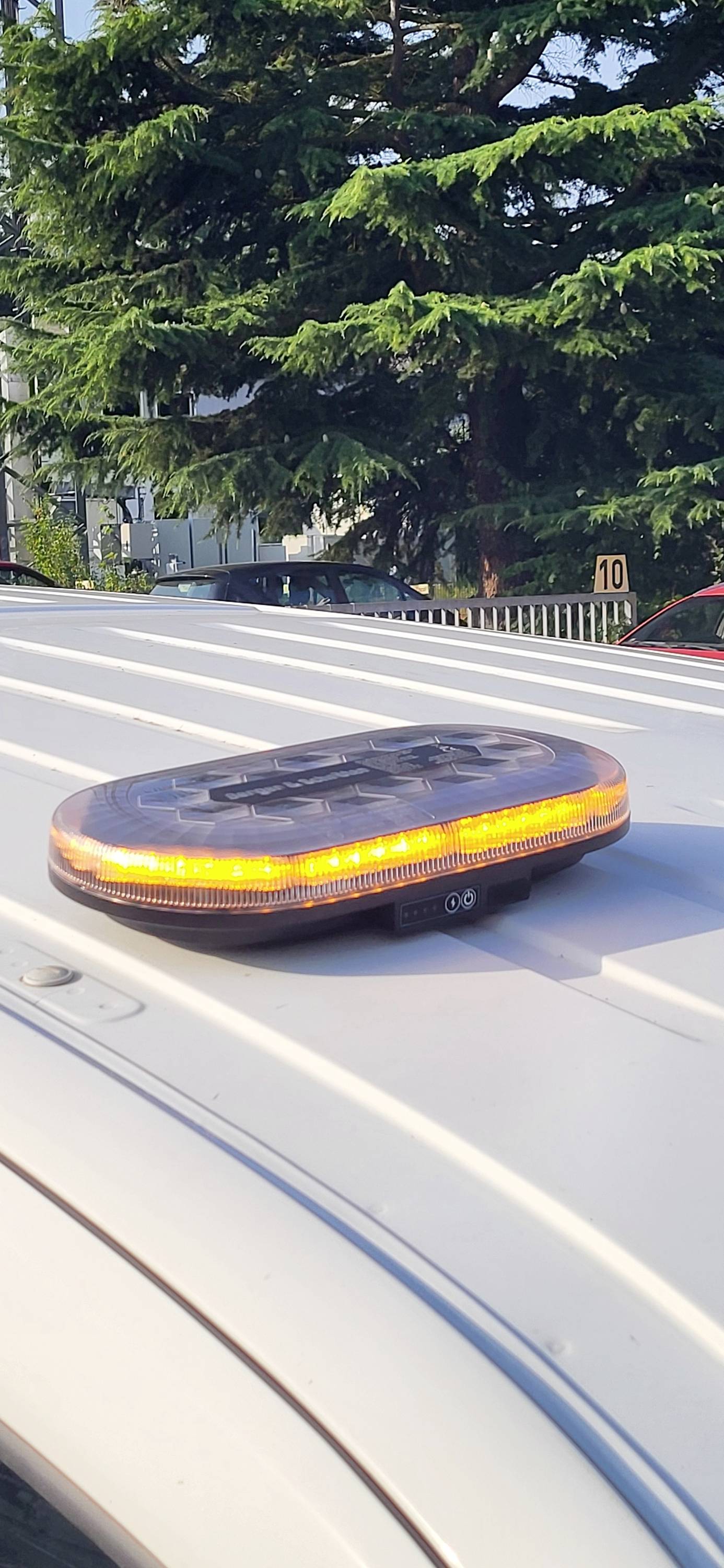 Magnetyczna lampa awaryjnego oświetlenia z bursztynowo-kolorową diodą LED jest zamontowana na dachu pojazdu. W tle widoczne są drzewa i znak drogowy.