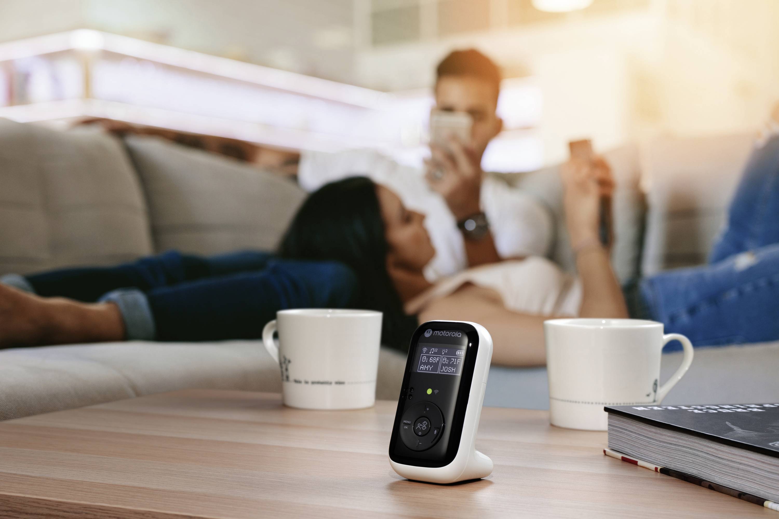 Monitor dla niemowląt leży na stole na pierwszym planie, obok dwóch kubków do kawy, podczas gdy para relaksuje się na kanapie w rozmytym tle.