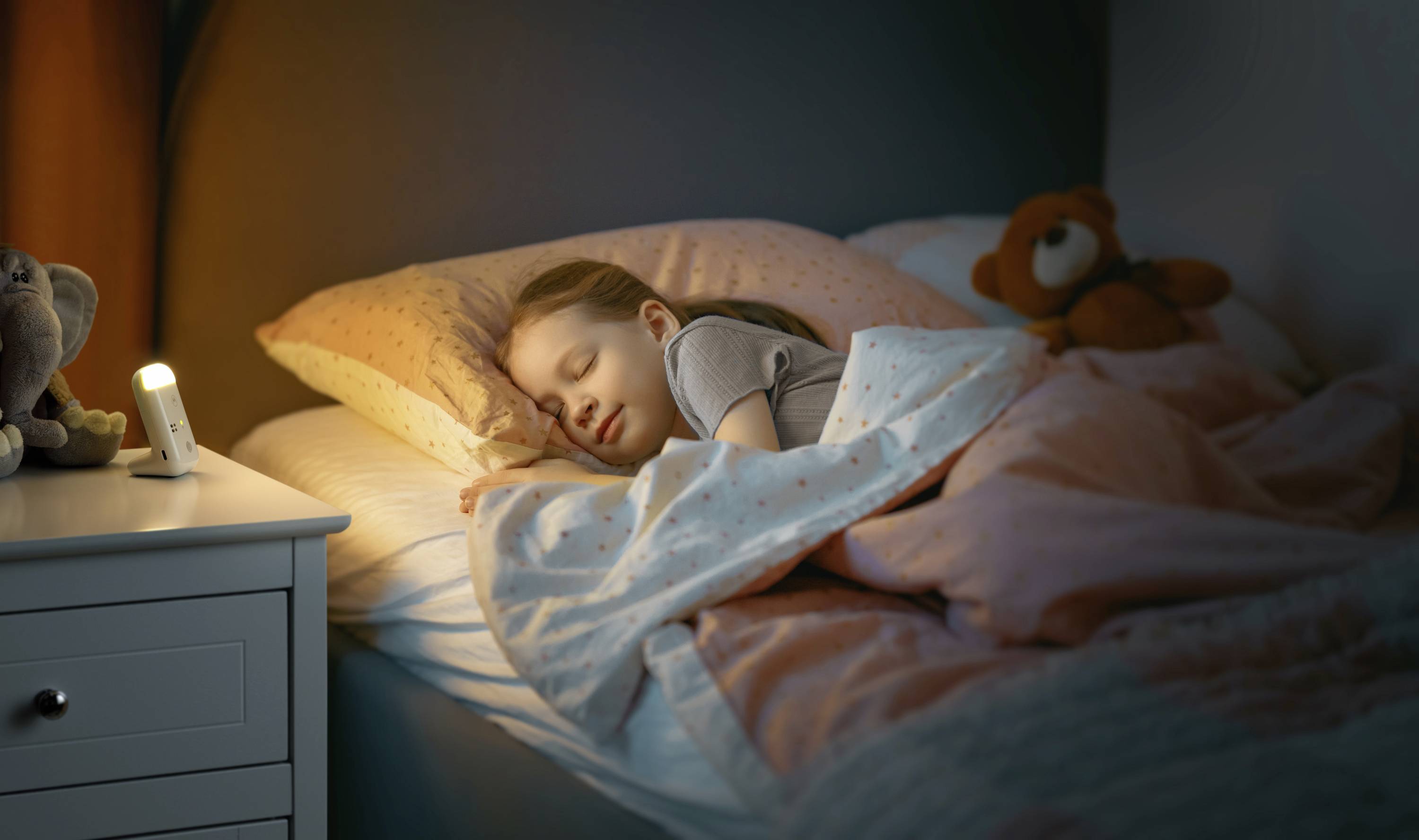 Dziecko spokojnie śpi w łóżku, a mała nocna lampka stoi na stoliku nocnym. Pluszowy miś leży obok poduszki.