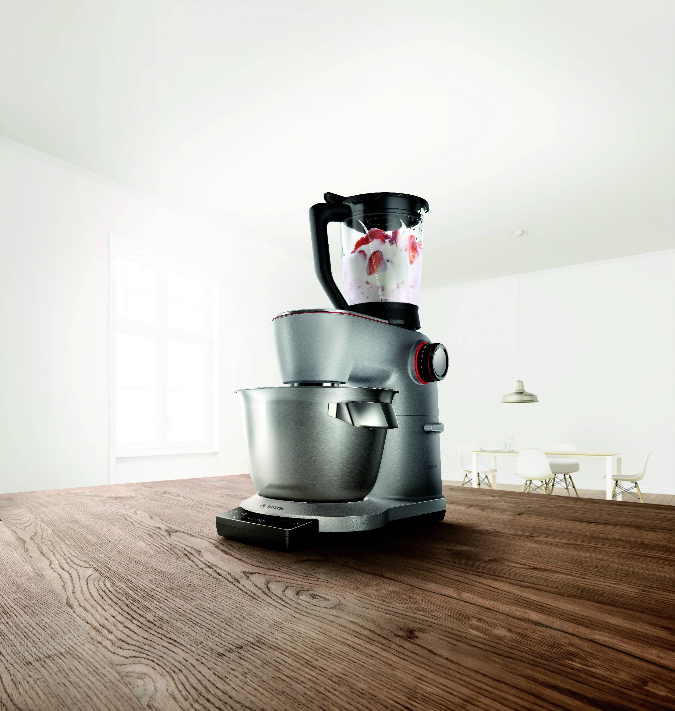 Srebrny mikser kuchenny z nasadką blendera na drewnianym blacie w jasnej, nowoczesnej kuchni z białymi ścianami i oknem.