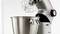 Srebrny robot kuchenny ze trzepaczką umieszczony jest nad dużą metalową miską, gotowy do użycia w przestrzeni kuchennej.