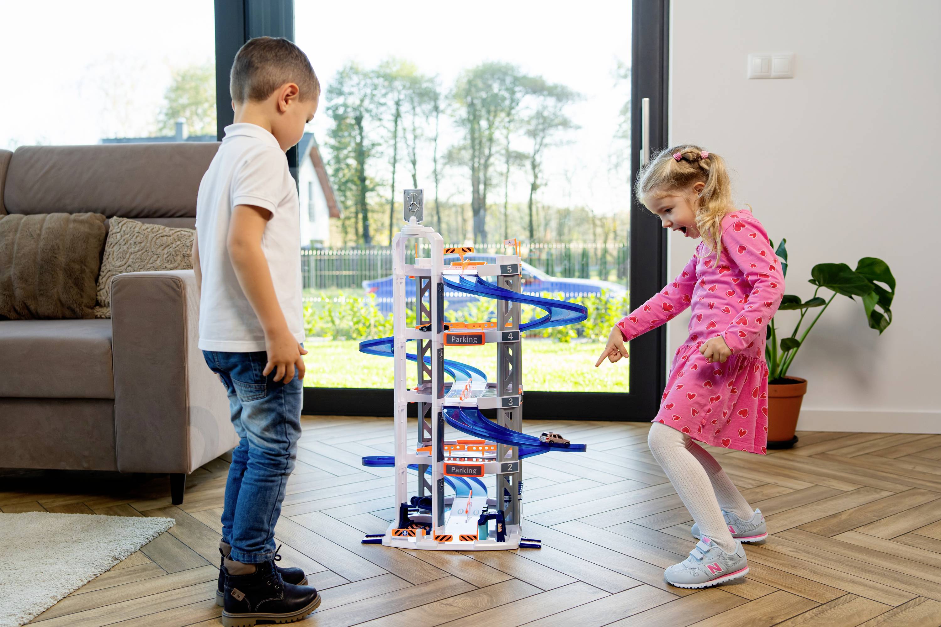 Chłopiec i dziewczynka bawią się wielopoziomowym torem wyścigowym w salonie, z dużymi oknami w tle, za którymi widać ogród.