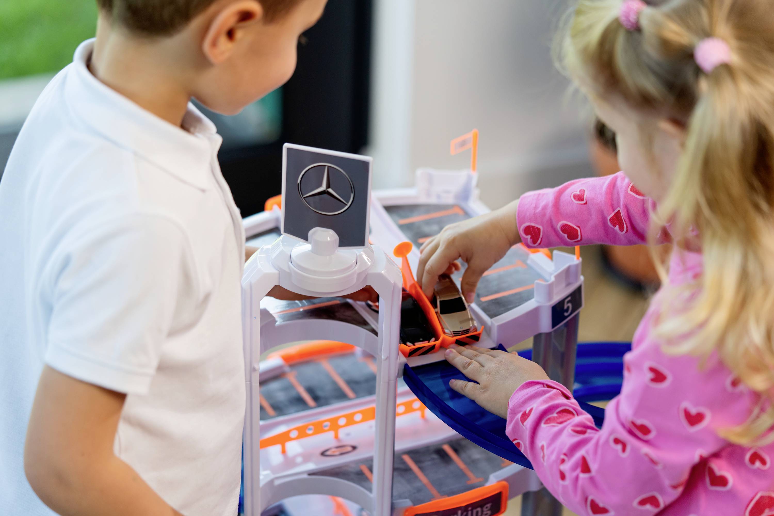 Dwoje dzieci bawiących się miniaturowym samochodem na wielopoziomowym zestawie do zabawy w postaci parkingu, z logo Mercedes, w pomieszczeniu.