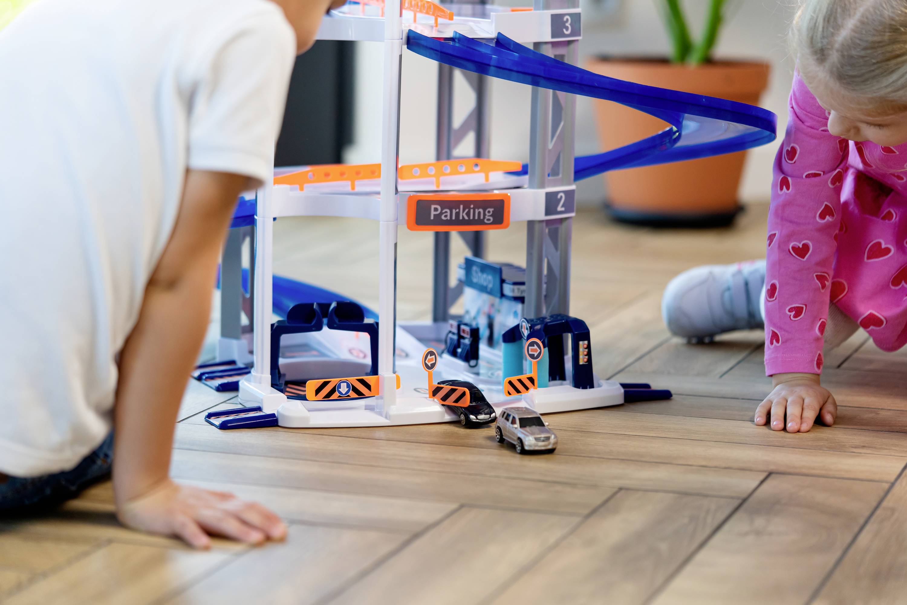 Dzieci bawiące się wielopoziomowym garażem zabawkowym na drewnianej podłodze; w zestawie wielopoziomowe rampy i miejsca parkingowe.