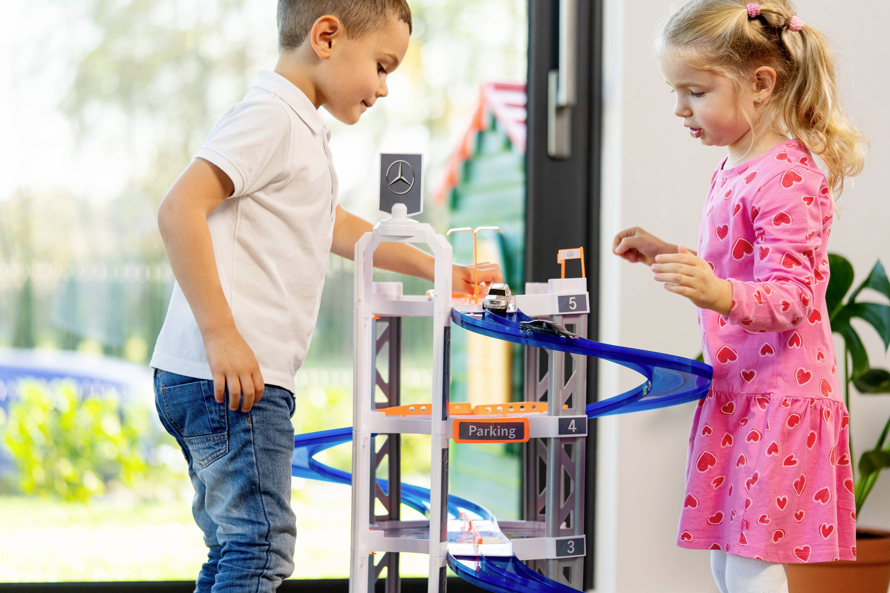 Chłopiec i dziewczynka bawią się zabawkowym parkingiem wielopoziomowym z niebieskimi zjazdami w jasno oświetlonym pomieszczeniu.