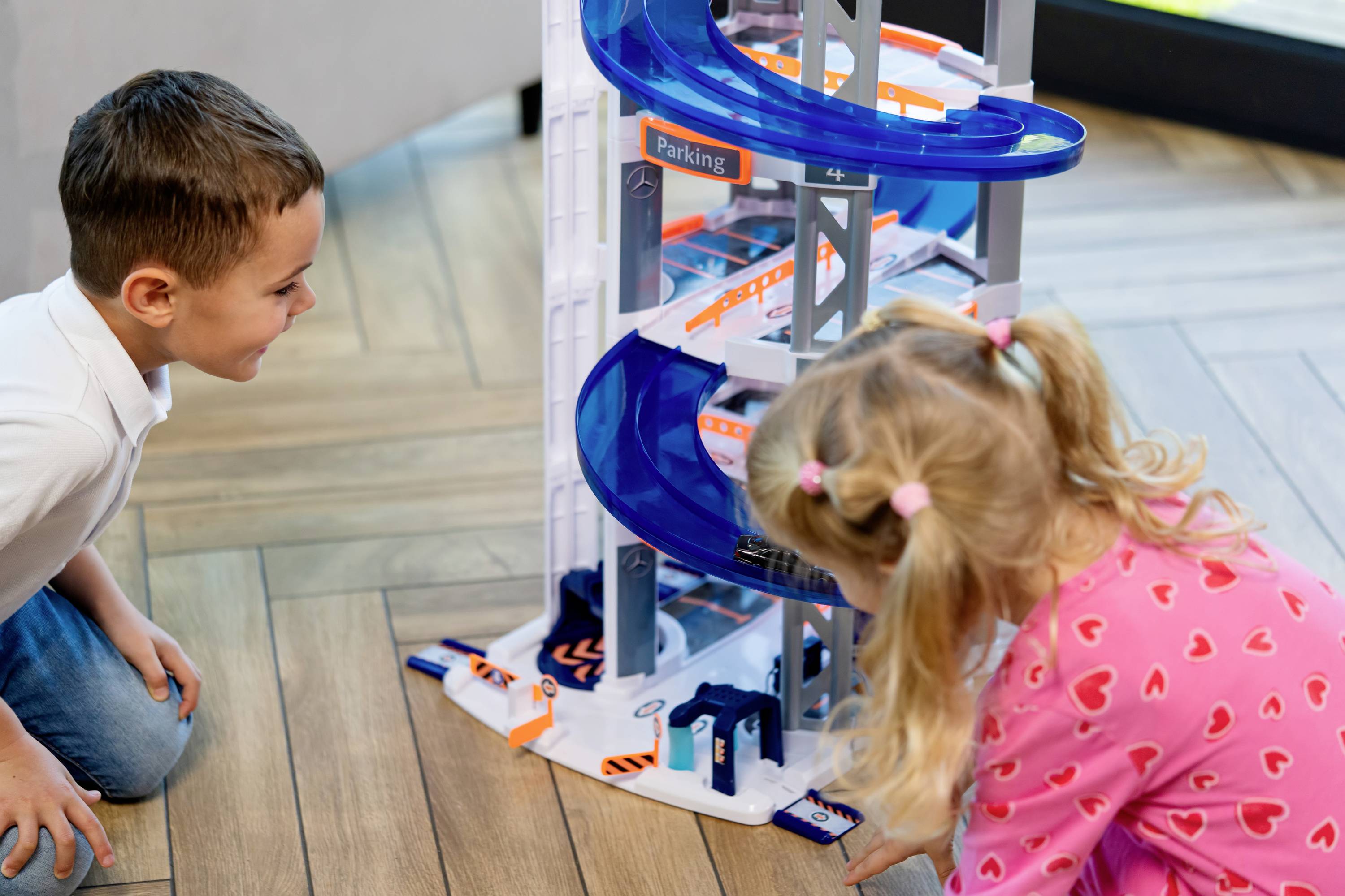 Dwoje dzieci bawi się wielopoziomowym parkingiem zabawkowym. Chłopiec się uśmiecha, podczas gdy dziewczynka z warkoczykami jest całkowicie skupiona na zabawie.