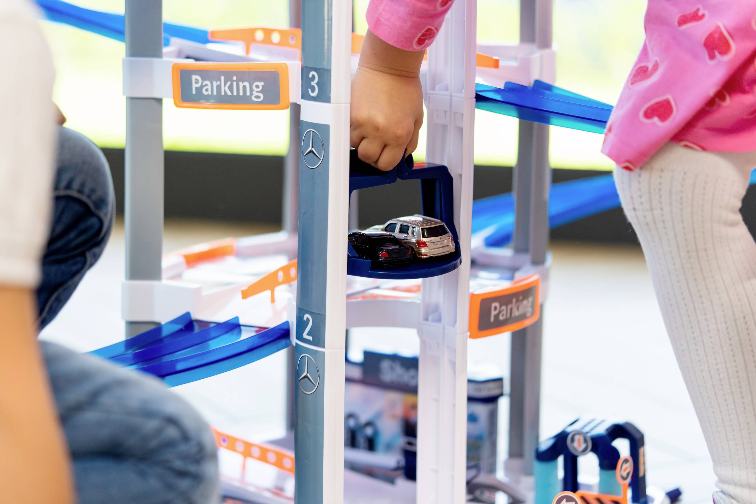 Dzieci bawią się miniaturowym parkingiem, który posiada wiele poziomów i pochylnie. Dziecko wkłada zabawkowy samochodzik do windy.