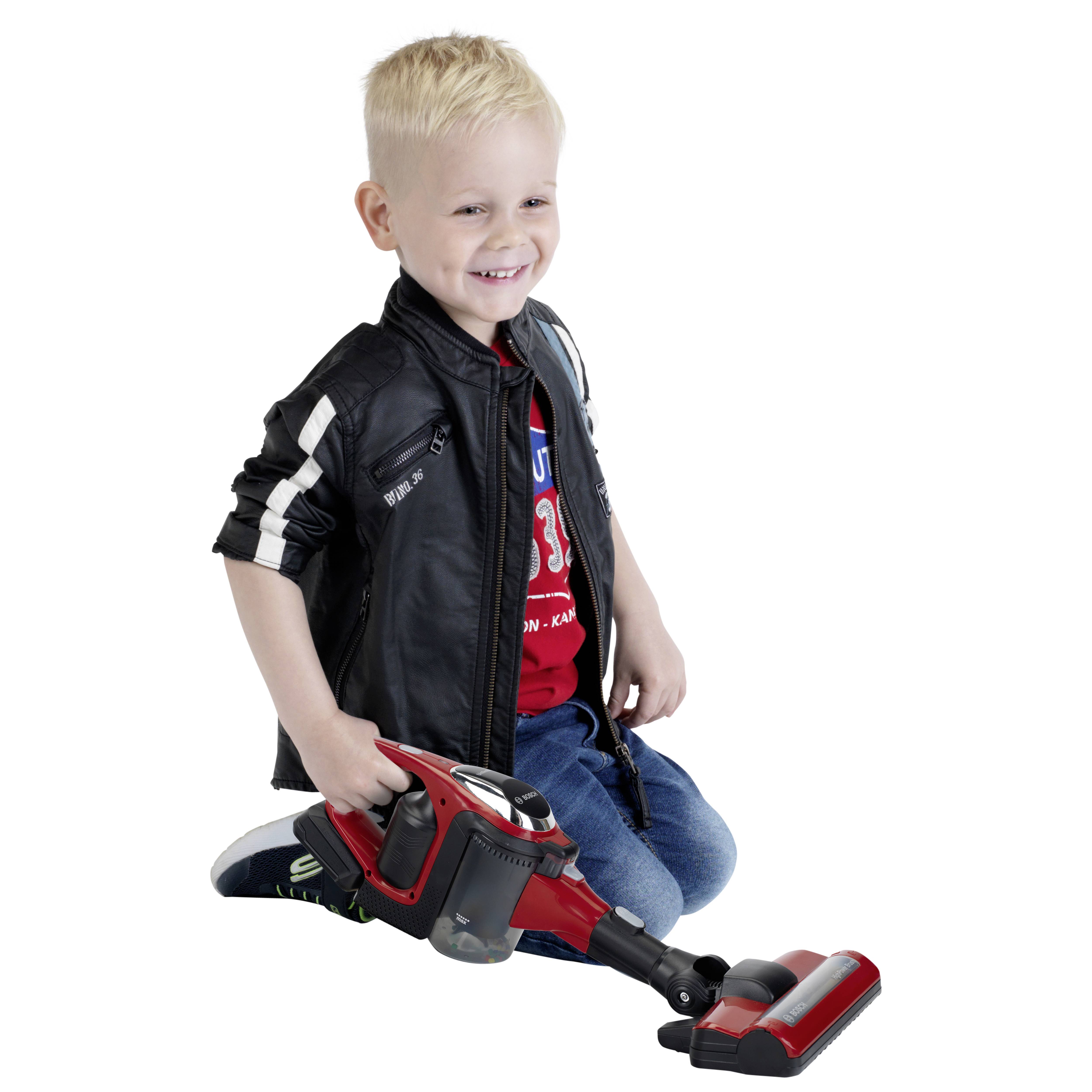 Młode dziecko o blond włosach uśmiecha się, trzymając zabawkowy odkurzacz na podłodze, ubrane w casualowe ubranie, w tym kurtkę.