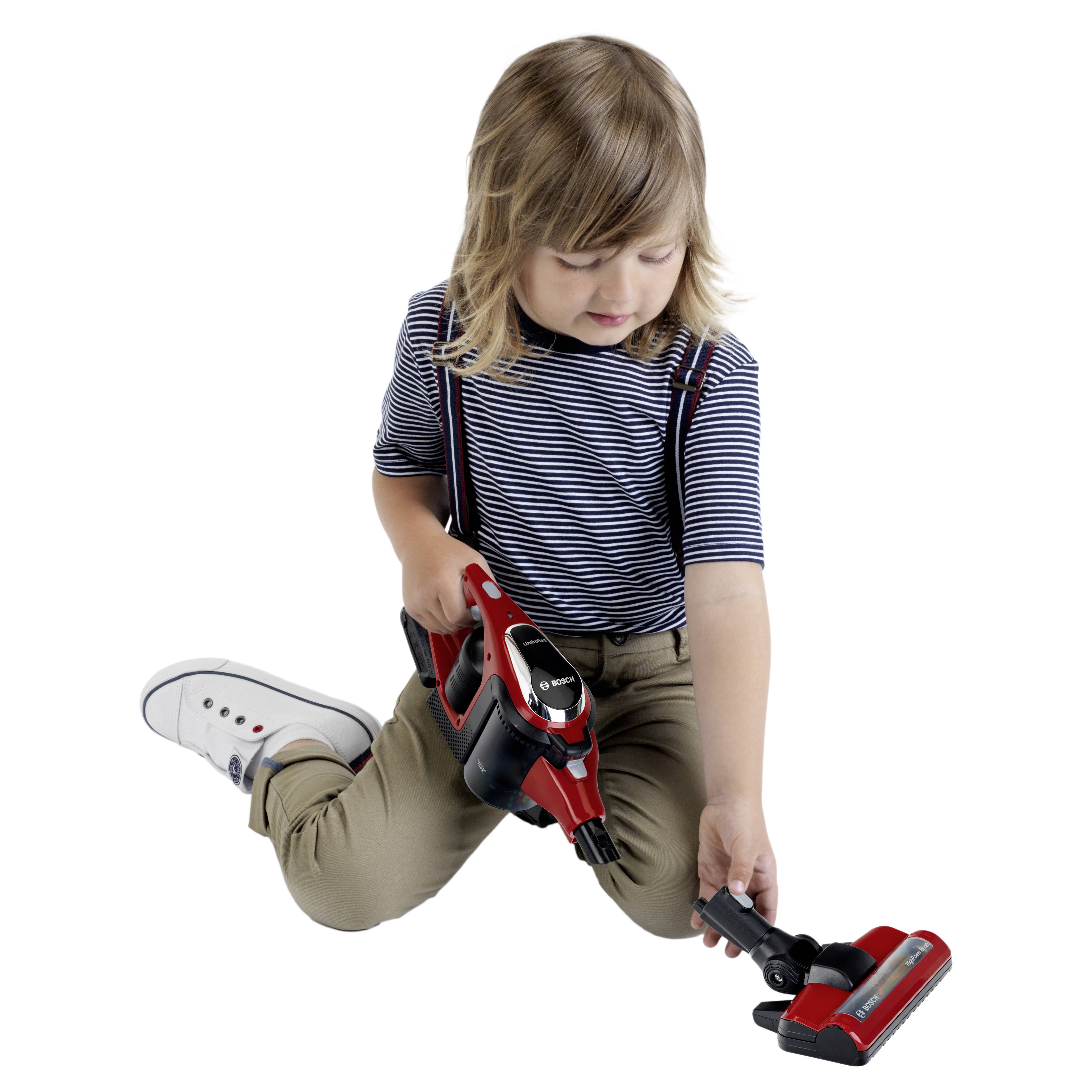 Dziecko siedzi na podłodze, trzymając i dokładnie oglądając czerwono-czarny odkurzacz z ciekawością, ubrane w pasiastą koszulkę i khaki spodnie.
