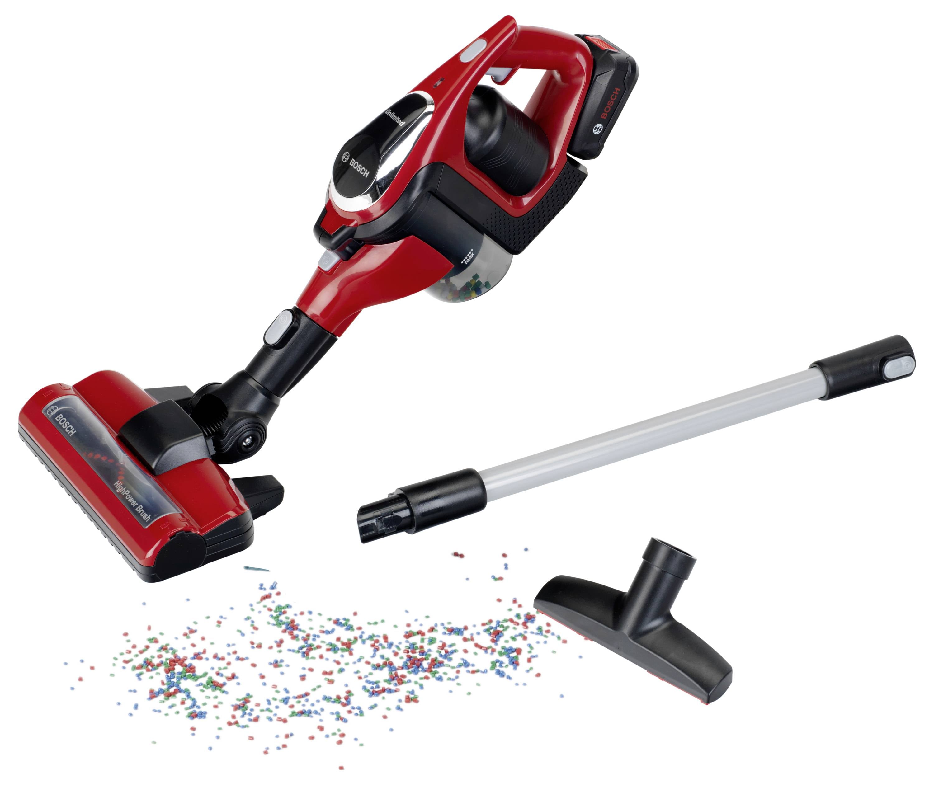 Czerwony i czarny bezprzewodowy odkurzacz stojący obok sterty kolorowych konfetti na podłodze, z dodatkowymi akcesoriami ułożonymi w pobliżu.