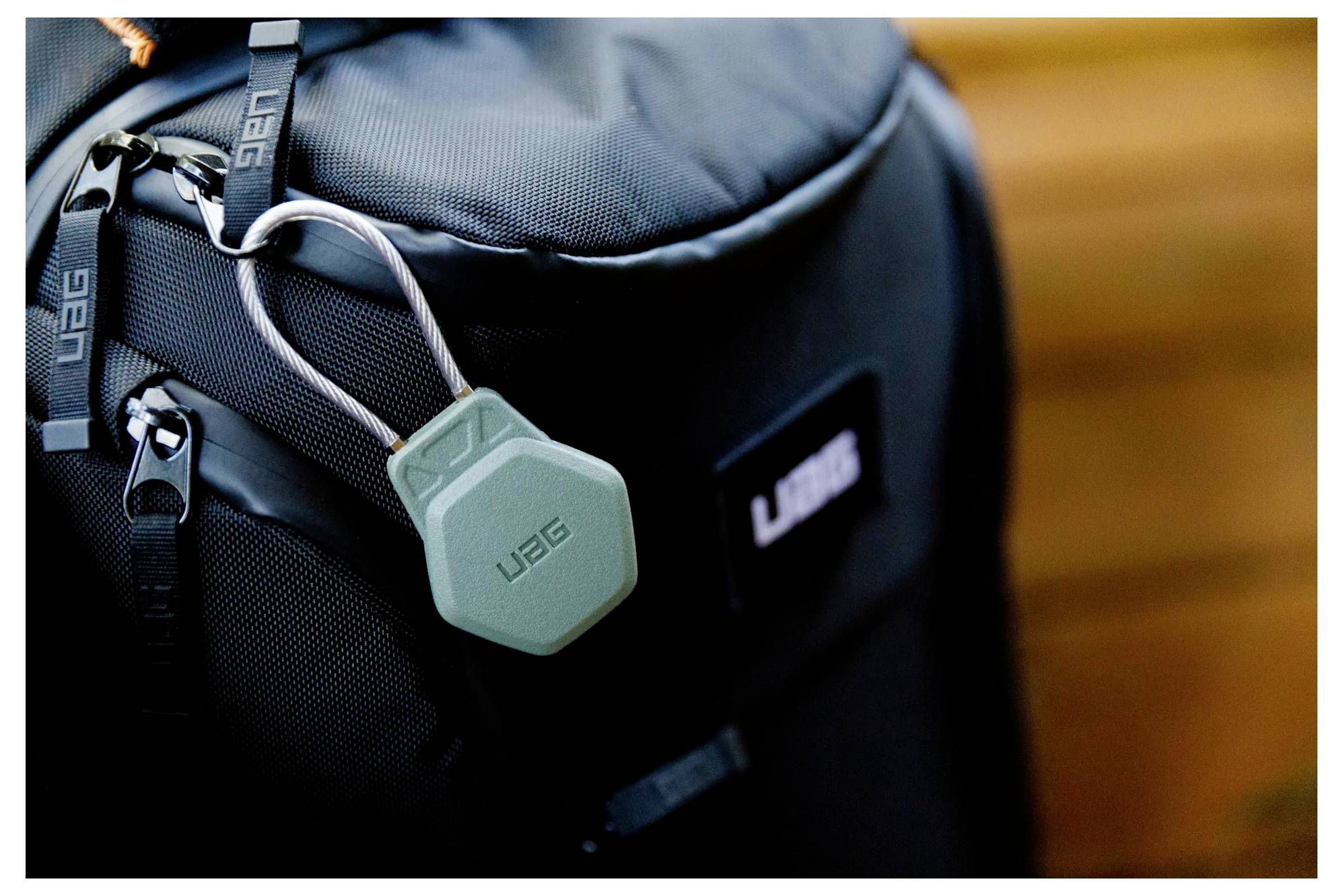Plecak posiada zieloną sześciokątną metkę z metalowym klipsem. Metka jest oznaczona logo 'UAG' i przymocowana do języka zamka błyskawicznego na torbie.