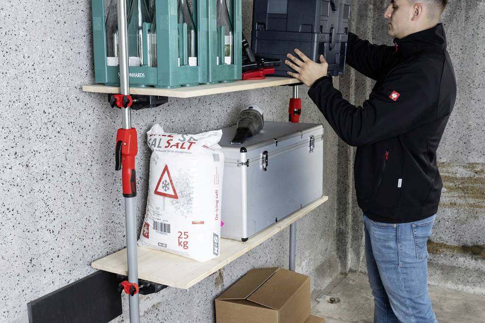 Osoba organizuje narzędzia i sprzęt na drewnianych półkach w garażu. Półki mieszczą zielone skrzynki narzędziowe, duży metalowy pojemnik oraz worek soli.