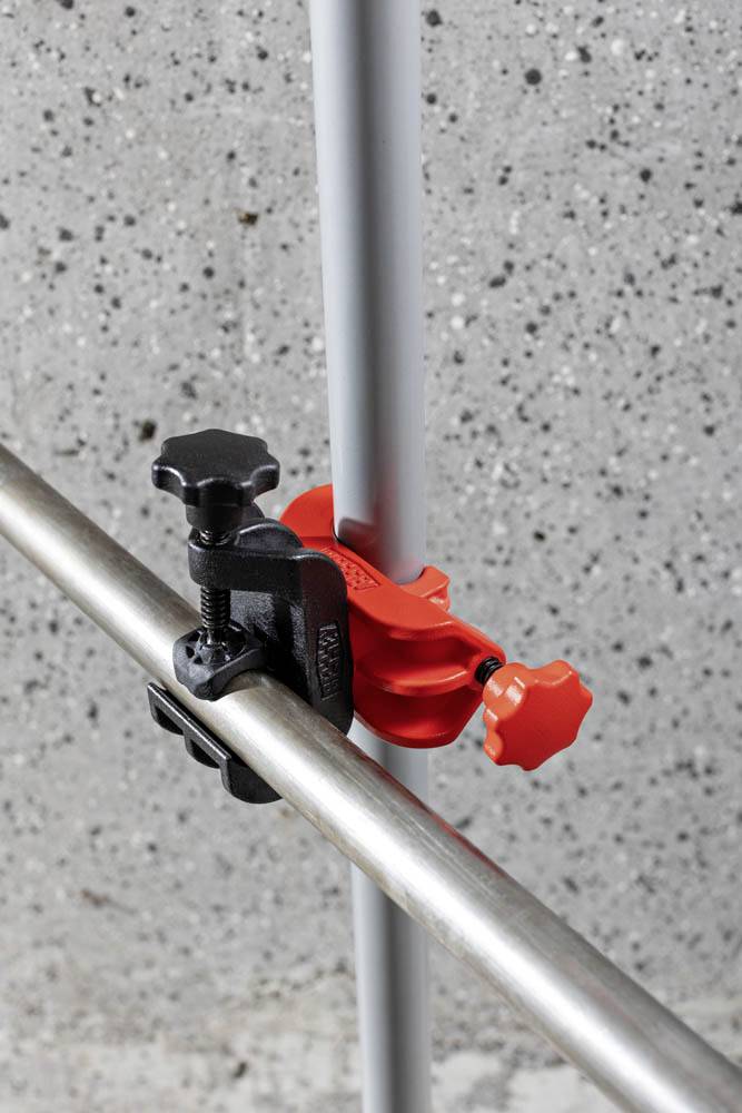Czerwono-czarny regulowany zacisk mocuje poziomą srebrną rurę metalową do rury pionowej na tle plamistej betonowej ściany.