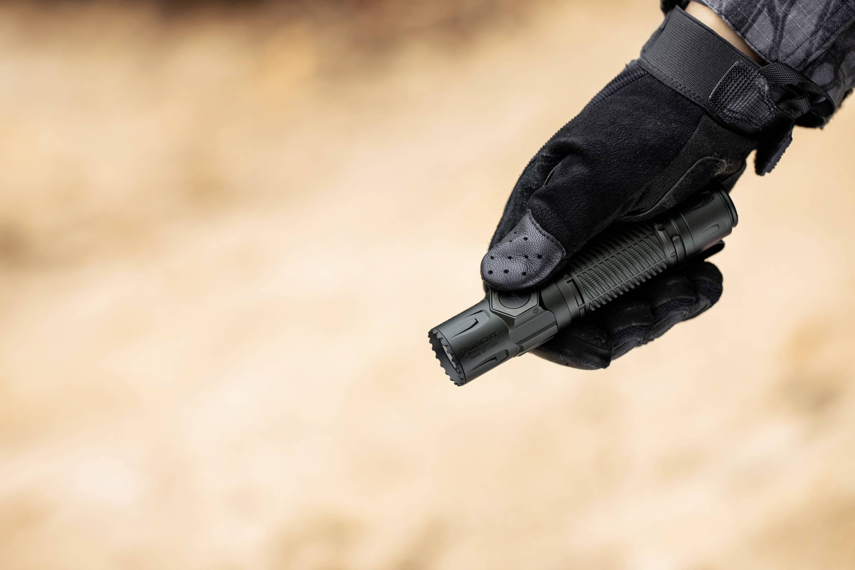 Rękawicowana dłoń trzyma czarną latarkę na rozmytym, brązowawym tle, sugerując użycie w terenie lub kontekst taktyczny.