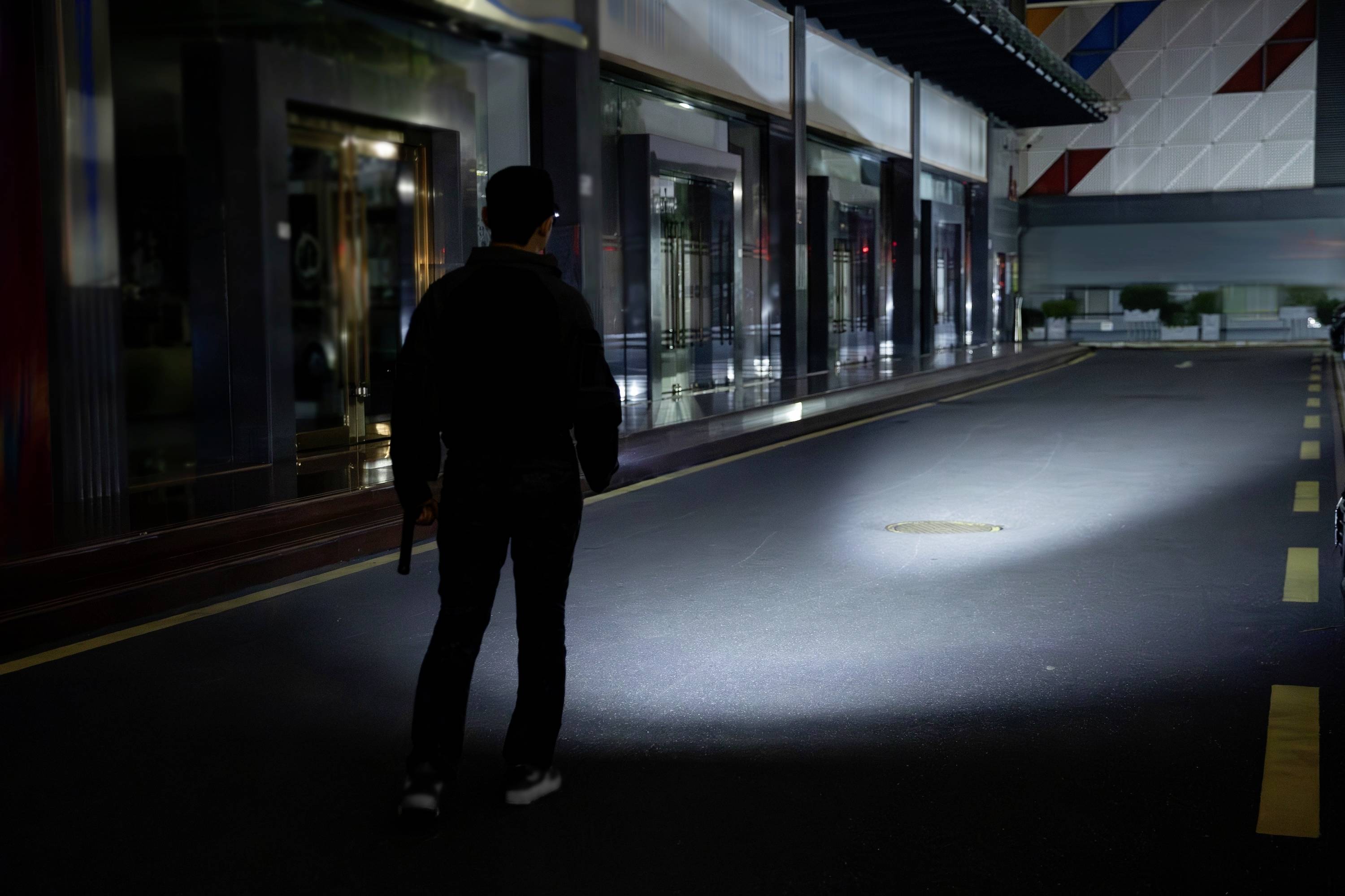 Osoba stoi na słabo oświetlonej ulicy, trzymając latarkę i oświetlając nią okrągłą plamę na ziemi. W tle widać budynki.