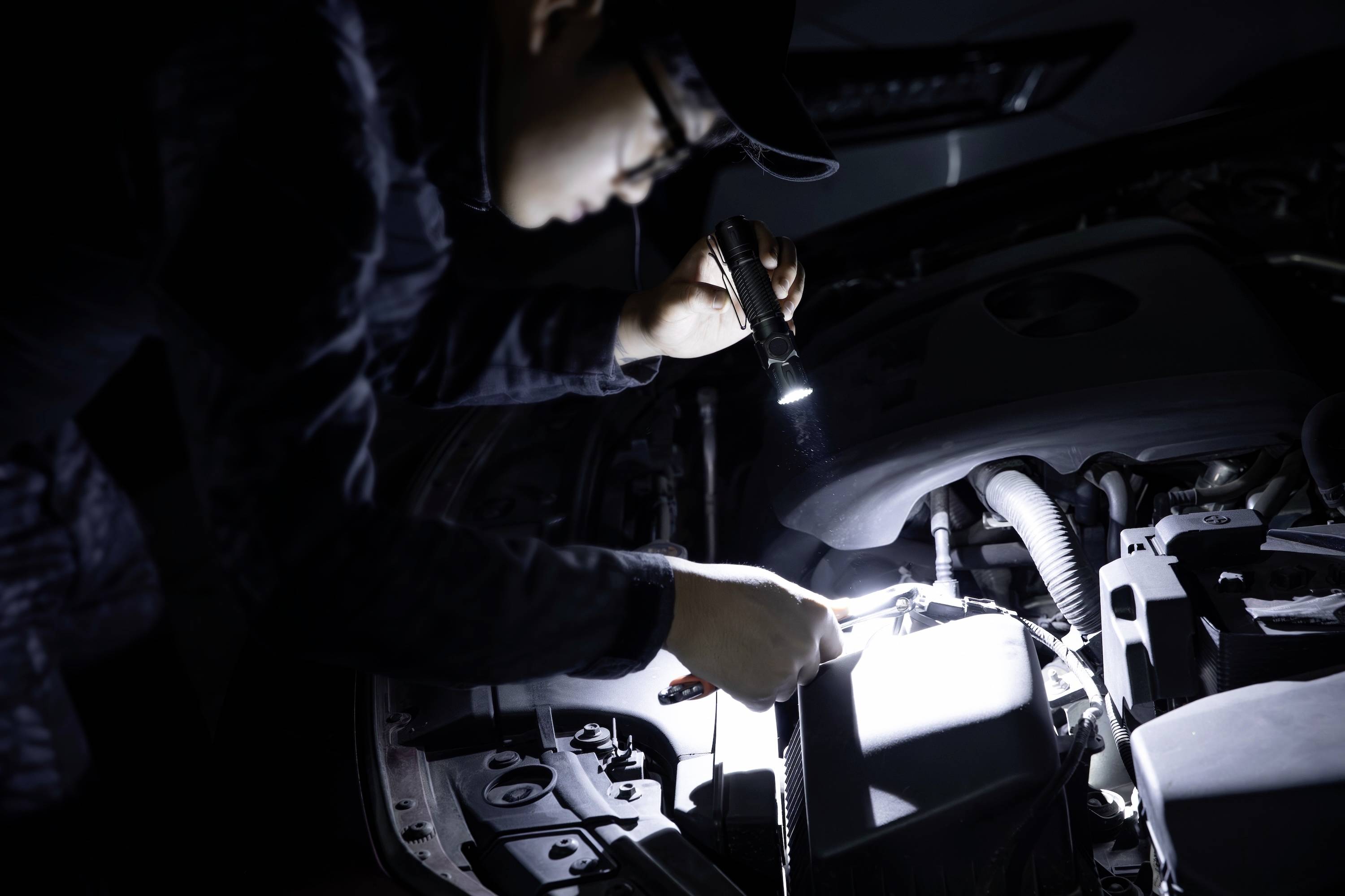 Osoba sprawdza silnik samochodu latarką w nocy, oświetlając poszczególne podzespoły.