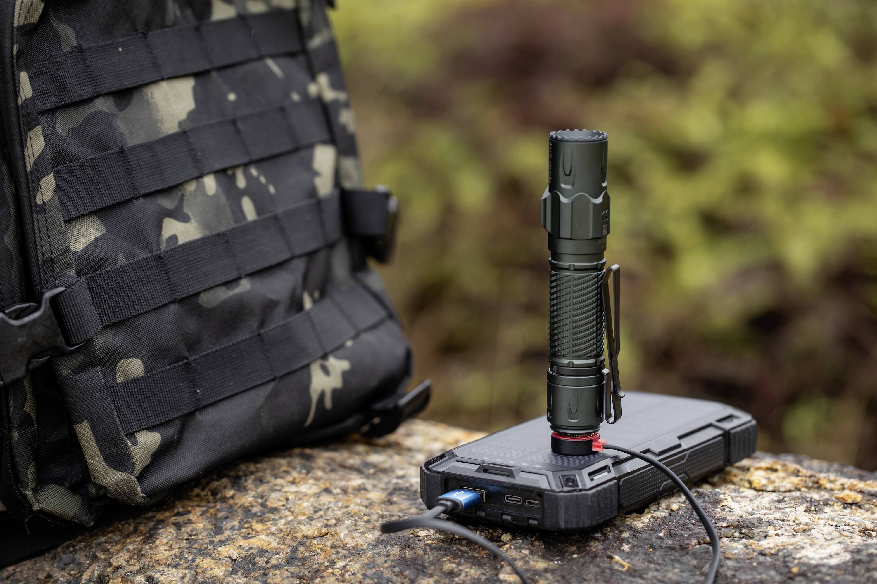 Taktyczna latarka ładująca się na powerbanku, leżącym na skale obok plecaka w kamuflażu, na wolnym powietrzu.