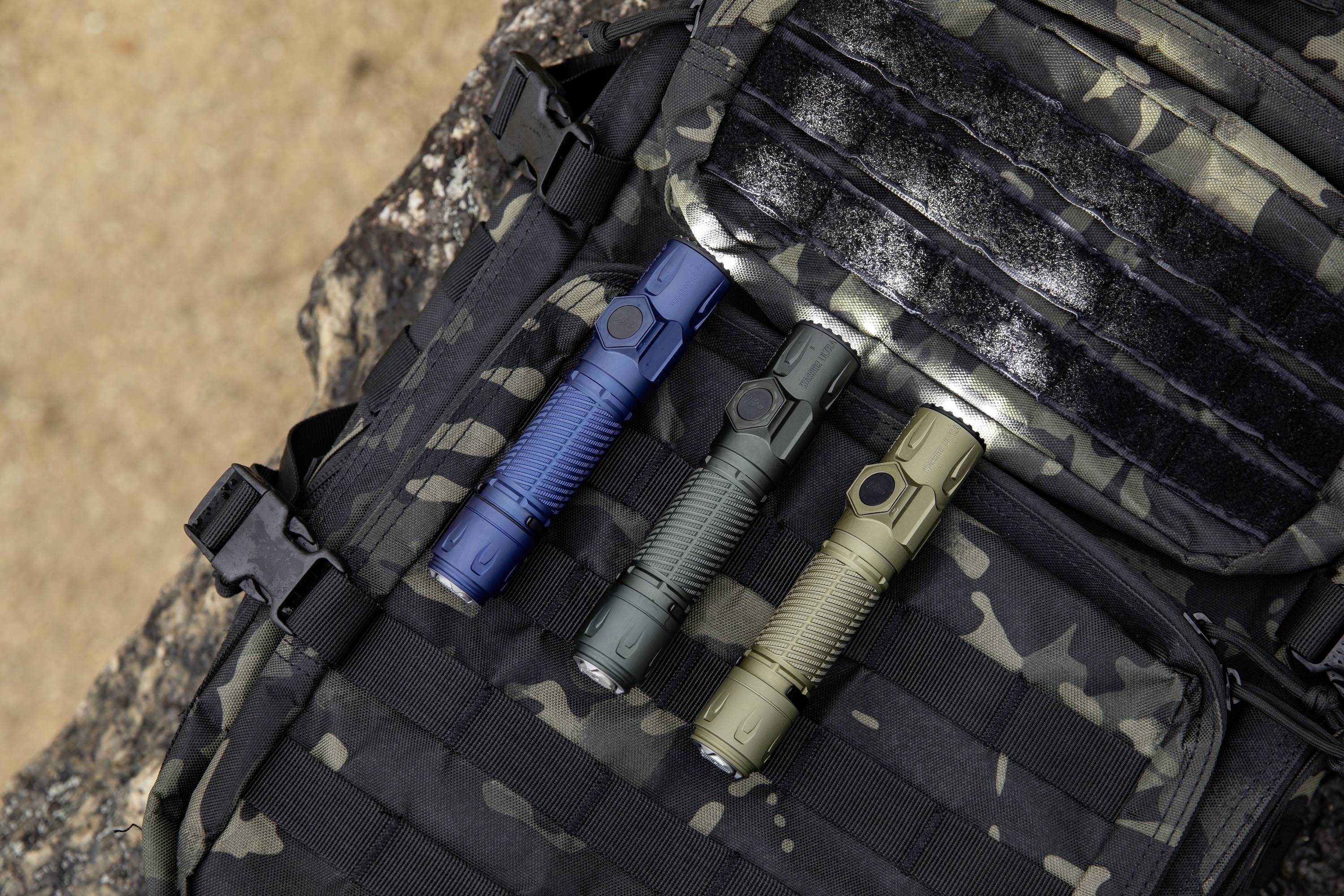Trzy taktyczne latarki, w kolorach niebieskim, czarnym i zielonym, leżą na plecaku we wzór maskujący, sugerując przeznaczenie outdoorowe lub taktyczne.