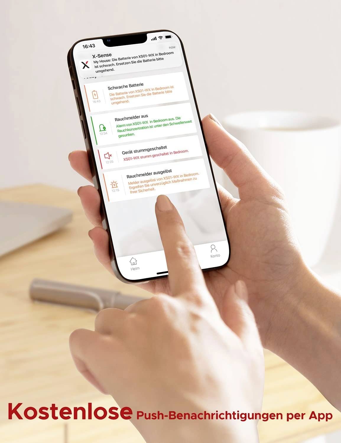 Dłoń trzymająca smartfon z aplikacją ostrzegającą o czujniku dymu. Ekran wyświetla powiadomienia dotyczące baterii, statusu czujnika i alarmów. Tło jest rozmyte.