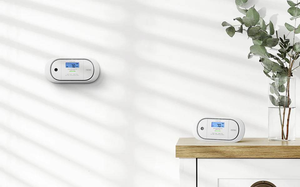 Zamontowany na ścianie monitor jakości powietrza wyświetla '72', inne urządzenie stoi na drewnianej półce. Słoneczne cienie padają na białą ścianę. Eukaliptus w wazonie.