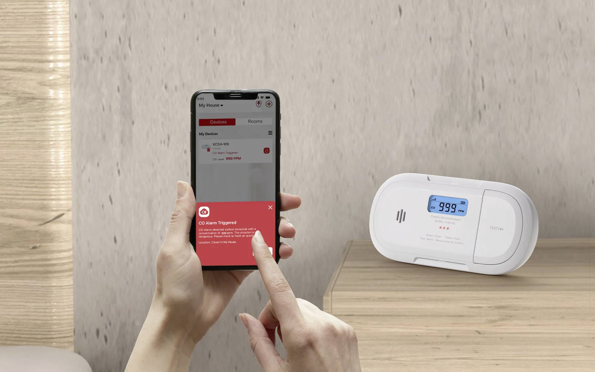 Komunikat "Alarm CO" wyświetlany na telefonie trzymanym przez osobę, obok cyfrowego detektora tlenku węgla wskazującego "999 ppm".